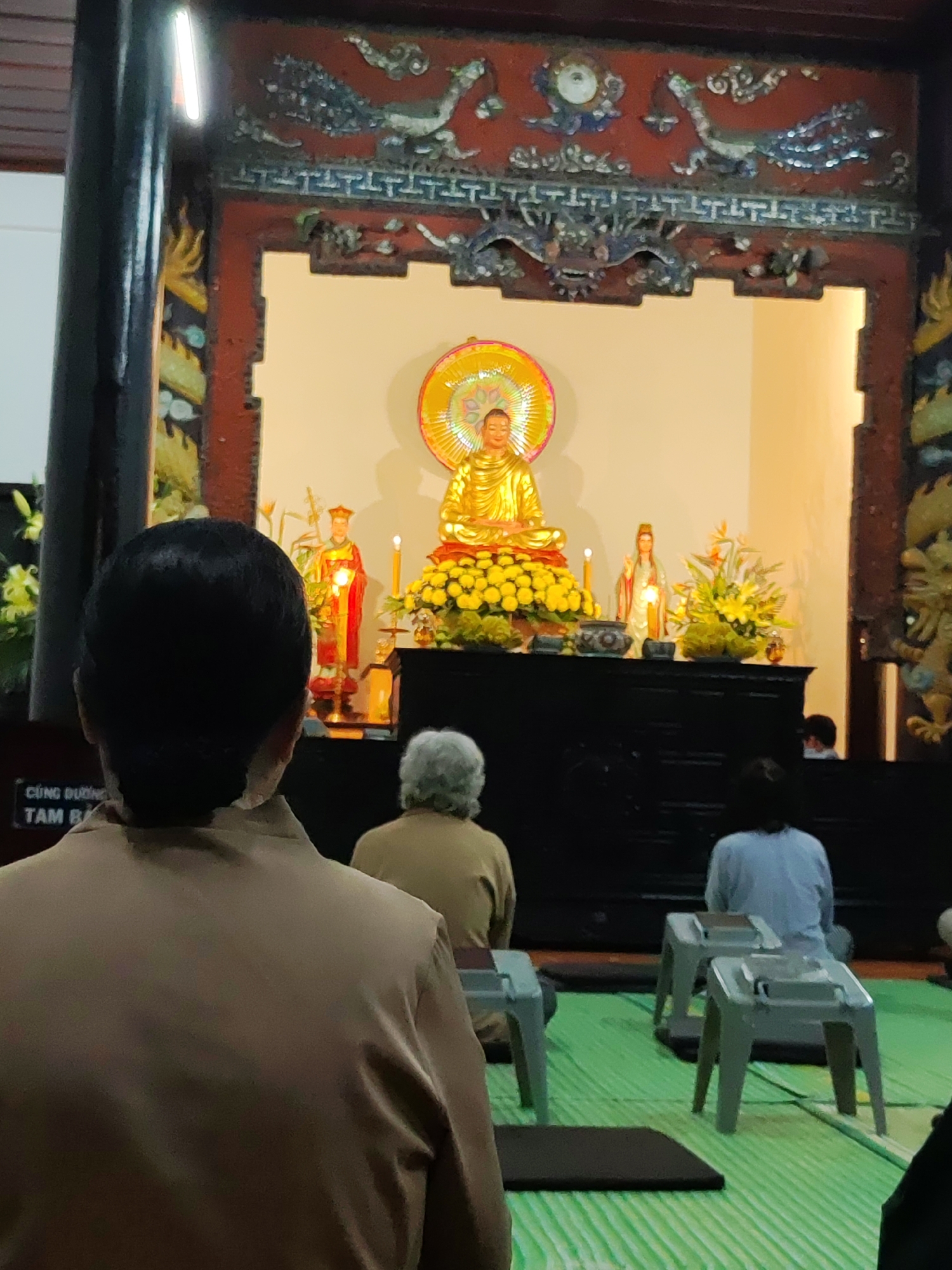 Captured in a temple in Da Lat, Vietnam, the image presents a gathering of people engaged in a meditative or devotional practice. In the foreground, the back of a person with dark hair and a brown shirt dominates the view, obscuring details beyond. Behind them, other people are seated on low stools on a green-striped floor. A black altar is adorned with a golden Buddha statue, flanked by figures of deities and floral arrangements, all illuminated by candlelight. The temple's architecture features intricate carvings on the surrounding frame, with phoenix and dragon motifs visible above the altar.