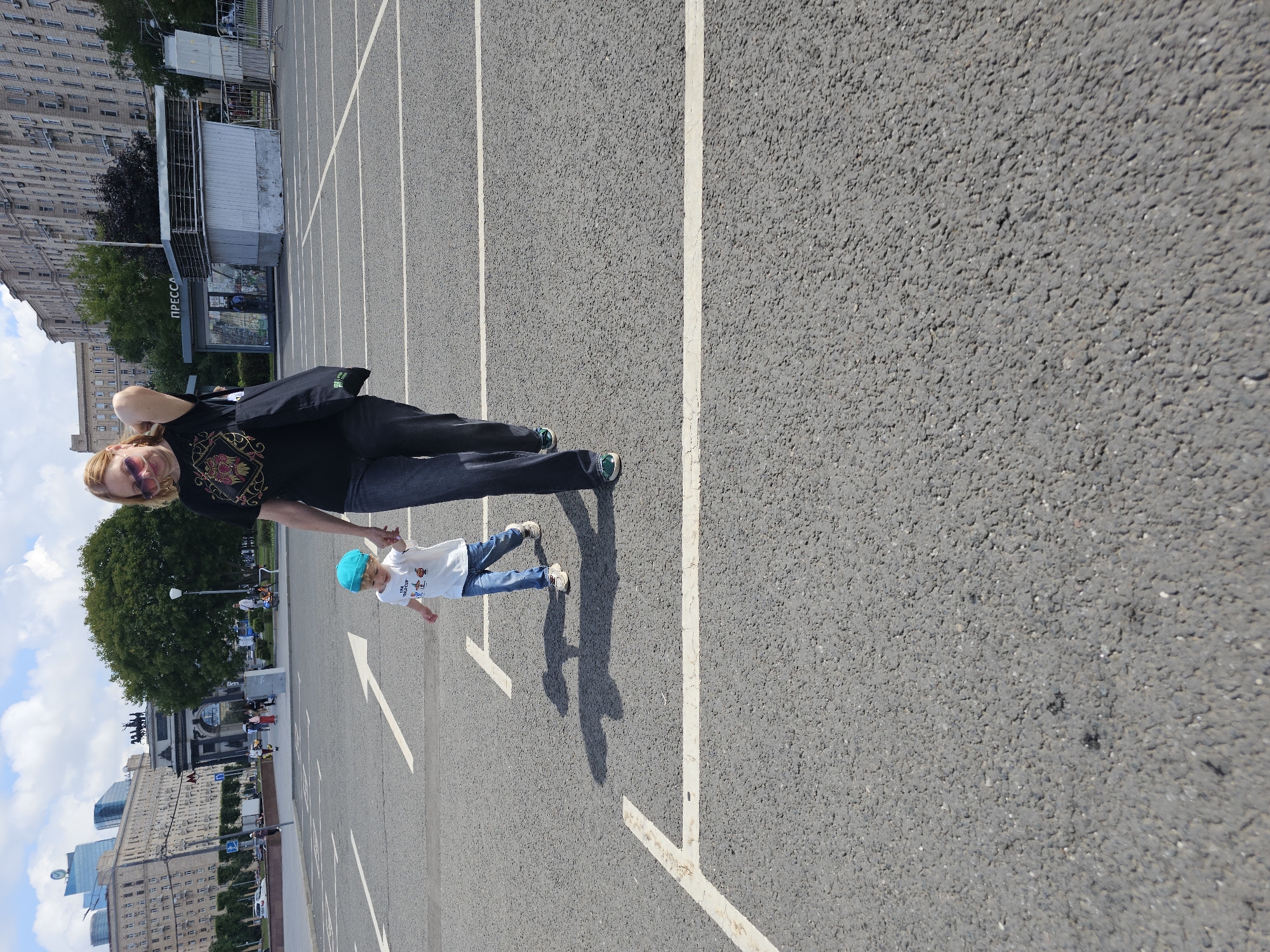 A woman in a black t-shirt and dark jeans walks with a young child wearing a blue hat and white t-shirt, holding the child's hand. They are on a paved surface marked with white lines, resembling a parking lot or a wide pedestrian area. In the background, there is a large, multi-story building, a triumphal arch with sculptures, trees, and a sky with scattered clouds. The environment suggests an urban setting in Moscow, Russia, potentially near a landmark given the presence of the arch and grand architecture. The woman carries a black tote bag over her shoulder. The child appears to be mid-stride.