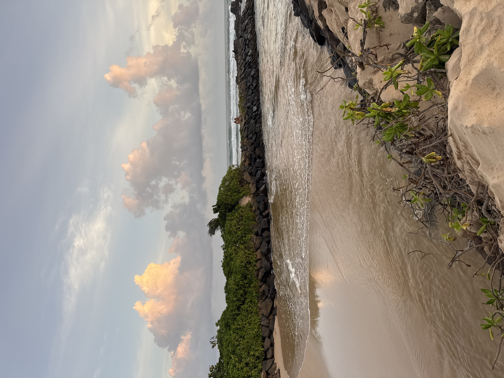 This image depicts a serene beach scene in Midigama, Sri Lanka. The composition is dominated by the tranquil ocean meeting a sandy shore, with a backdrop of dramatic clouds. In the mid-ground, a line of dark rocks forms a breakwater, separating the calmer water from the incoming waves. Two figures are visible sitting on this breakwater, appearing relaxed and enjoying the scenery.
The sky is a canvas of soft blues and grays, punctuated by large, billowy clouds that are tinged with warm, peachy hues, suggesting either sunrise or sunset. The weather appears calm, with gentle waves lapping at the shore. Lush green vegetation covers the higher ground behind the breakwater, adding a touch of vibrant color to the scene. The sandy beach stretches out to the right, reflecting the soft light from the sky. Some dry branches and green leaves are growing out of the sandy embankment on the right, indicating a natural, coastal environment.
The overall mood of the image is peaceful and idyllic, capturing a moment of quiet enjoyment of nature. There is no visible text in the image.