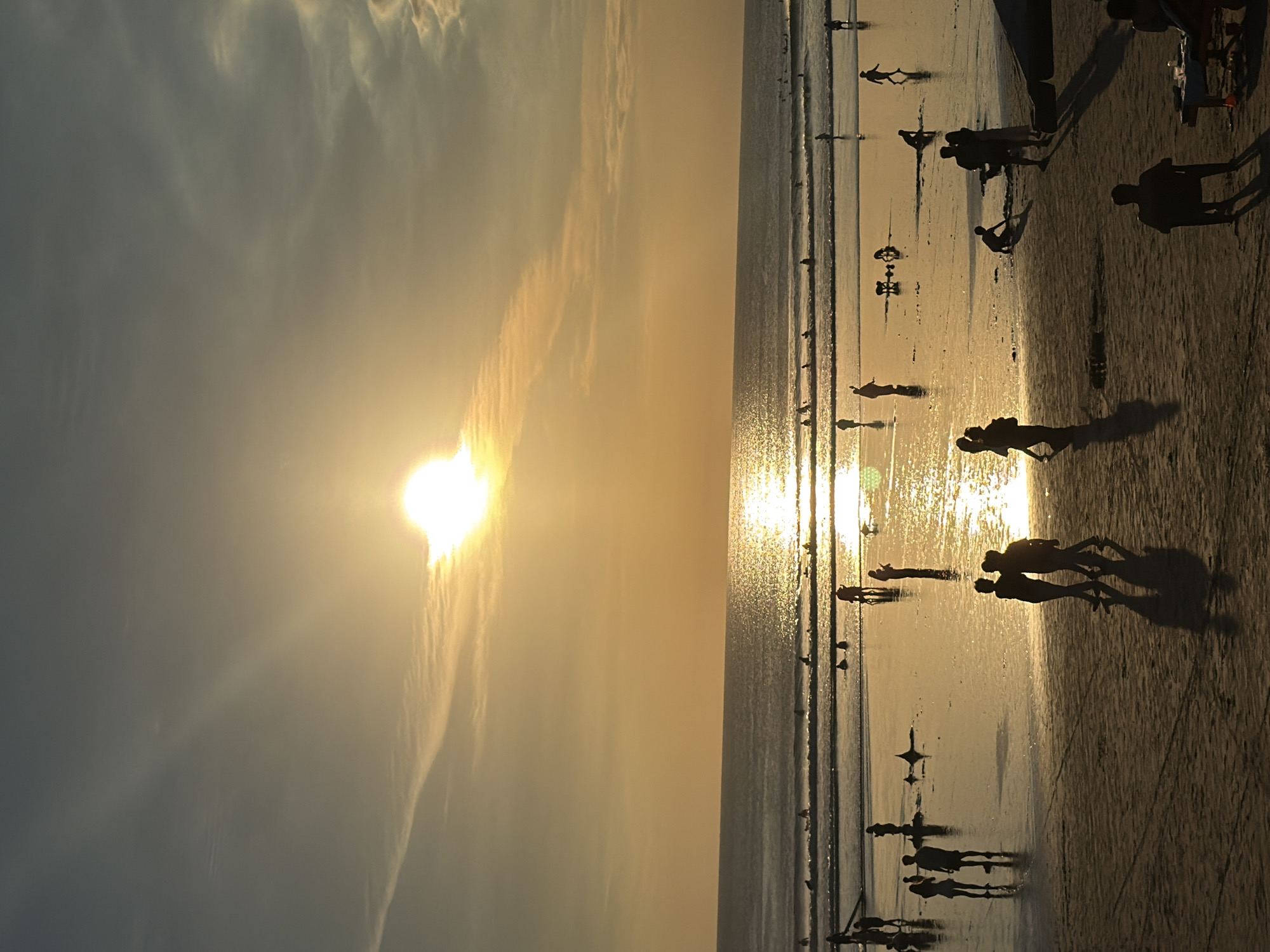 This is a photograph of Kuta Beach in Bali, Indonesia, at sunset. The sun is low on the horizon, casting a warm, golden light across the sky and water. The ocean is calm, with gentle waves rolling in. Several surfers are visible in the water, enjoying the last light of the day. On the beach, a few people are walking and playing, their silhouettes dark against the bright sand. The scene is peaceful and serene, capturing the beauty of a tropical sunset.