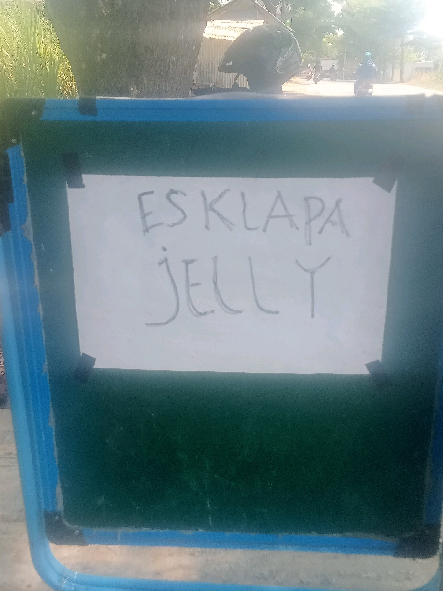 A whiteboard easel is placed outdoors on a paved surface, likely a roadside. The easel has a blue frame and a dark green writing surface. A white sheet of paper, attached with black tape, is affixed to the easel, displaying handwritten text in gray marker. The top line reads "ESKLAPA" and the bottom line reads "JELLY". In the background, a dirt road is visible with several motorcycles and people. A large tree trunk is prominent to the left of the road, and buildings with corrugated roofs are visible further back. The overall lighting suggests it is daytime. This scene is located in Penusupan, Indonesia.