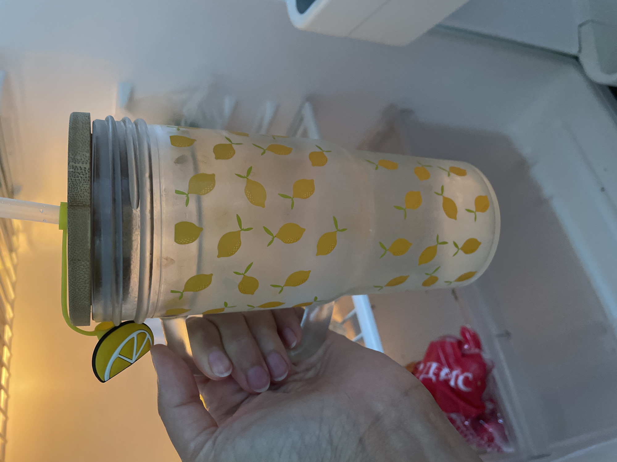 A hand is holding a glass tumbler with a lemon pattern and a bamboo lid, which is inside a refrigerator. The tumbler has a straw inserted through the lid and a small lemon-shaped charm attached to the straw. A red plastic package is visible on a shelf below the tumbler. The background of the refrigerator is illuminated by a yellow light, and condensation is visible on the interior walls.