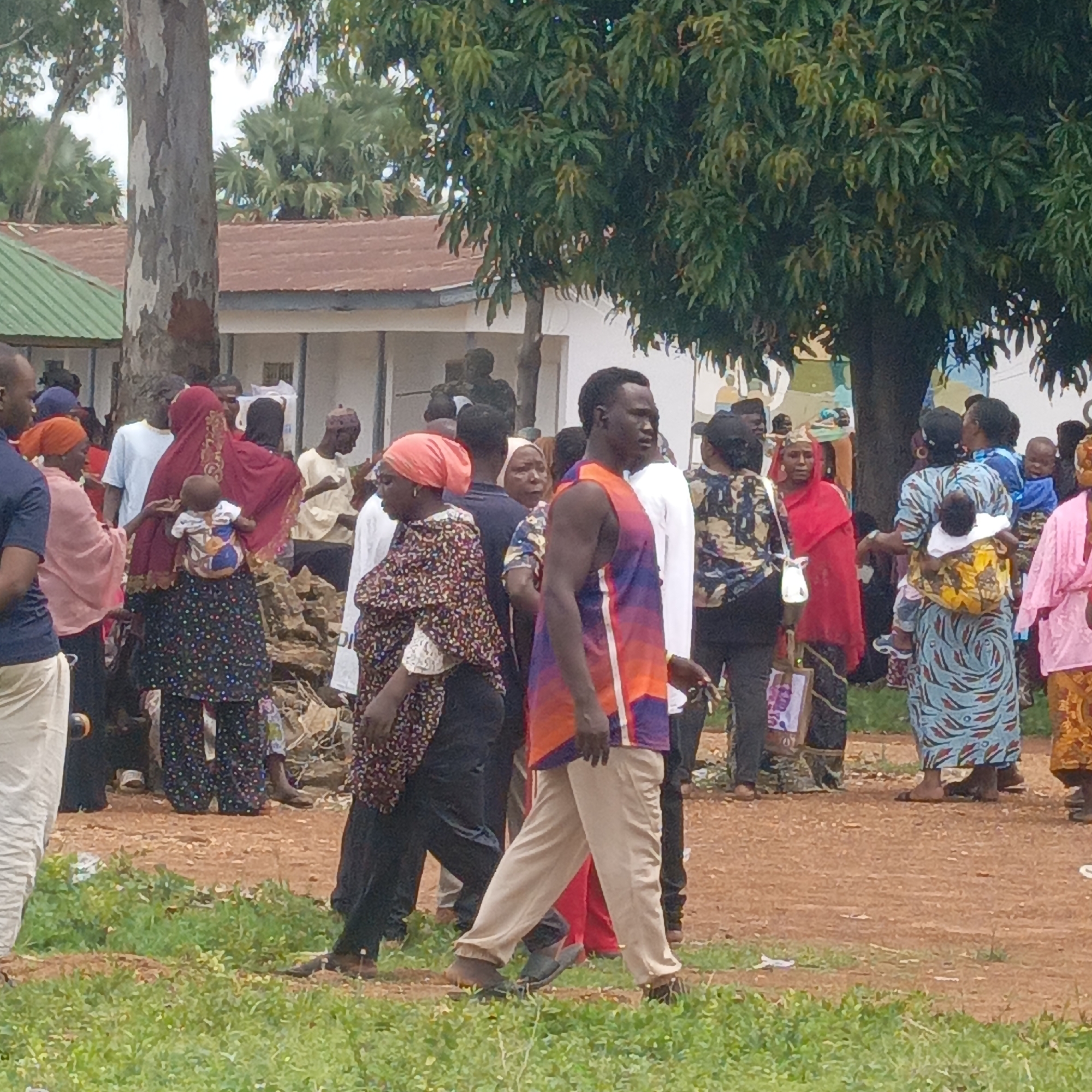 The image depicts a gathering of people in Akwanga, Nigeria. Predominantly women are present, many carrying babies on their backs. They are dressed in a variety of traditional African clothing, including headwraps and brightly colored fabrics. A man in a purple and orange tank top and khaki pants is walking through the crowd. The environment includes a building with a green and brown roof, trees, and dirt ground with patches of grass.