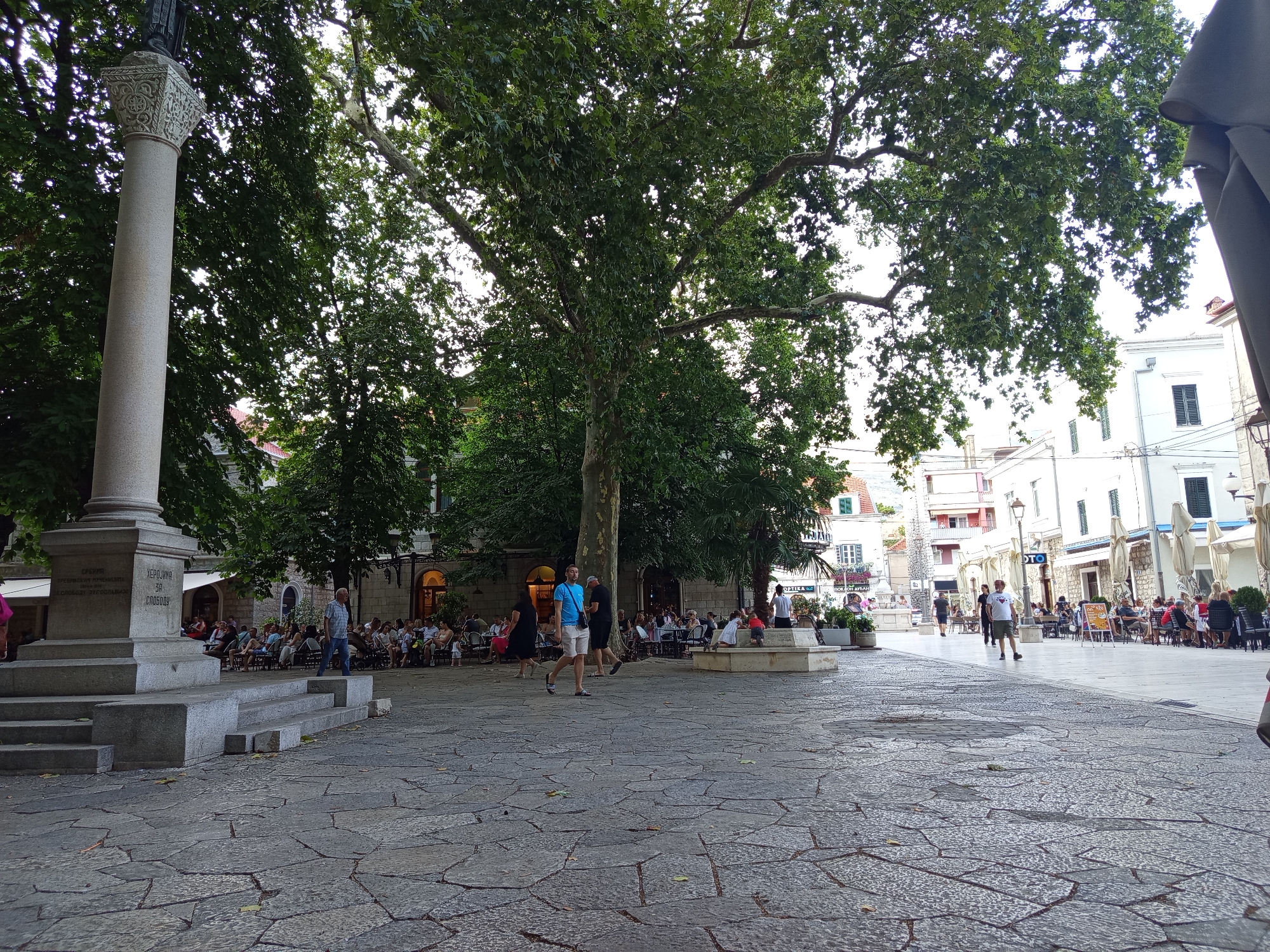 The image shows a daytime scene in Trebinje, Bosnia and Herzegovina, likely in the central square or a pedestrianized area. A large stone monument with carved inscriptions stands prominently on the left. In front of the monument, there are tiered stone steps leading up to it. The ground is paved with irregular flagstones.
Several people are visible throughout the scene. In the background, many individuals are seated at tables outside cafes or restaurants, appearing to be socializing or dining. Some people are walking across the square, while others are standing near the monument or interacting with each other. The area is shaded by large, mature trees with lush green foliage, casting dappled light on the ground. Buildings with awnings and outdoor seating line the perimeter of the square, indicating a commercial and social hub. The overall atmosphere suggests a lively public space during daylight hours.