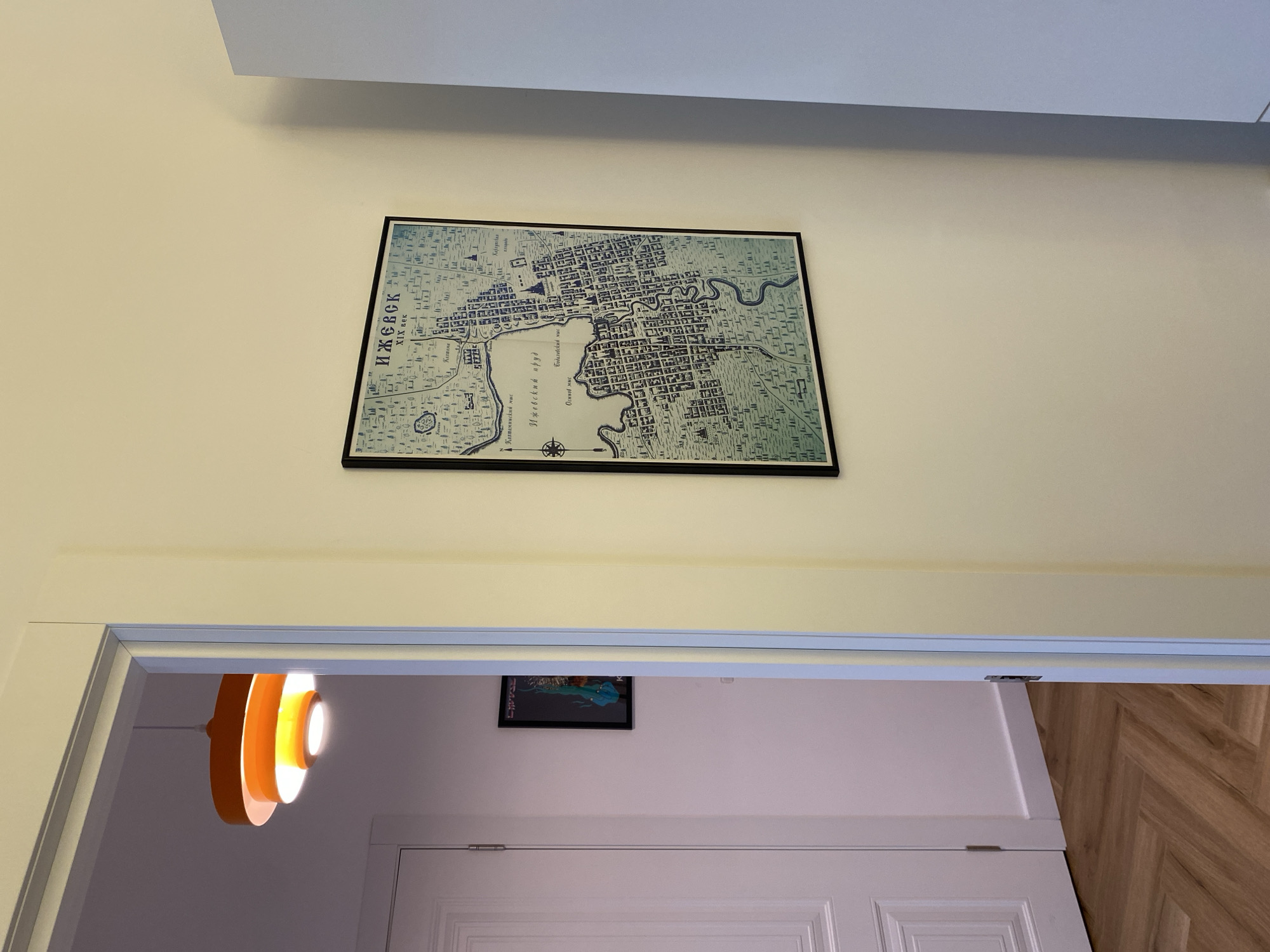The image shows an interior hallway with a light yellow wall on the right and a cream-colored wall on the left, visible through an open doorway. A framed map of Izhevsk, Russia, titled "ИЖЕВСК XIX век" (Izhevsk 19th century), is hung on the yellow wall. The map depicts the city's layout with buildings and streets. A portion of a white door and a herringbone-patterned wooden floor are visible through the doorway. An orange pendant light fixture hangs from the ceiling in the adjacent room. A framed artwork is also visible on the cream-colored wall.
