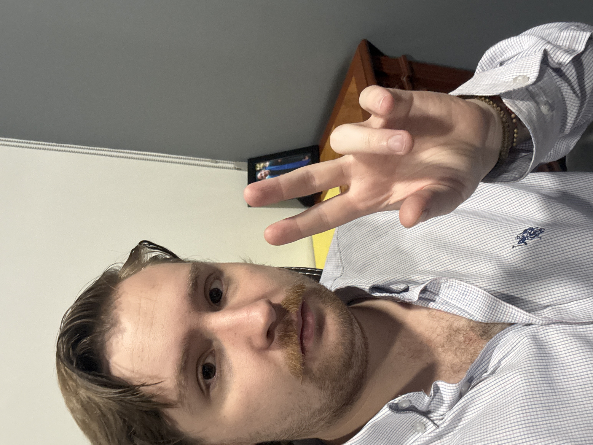 A man with light brown hair and a mustache is taking a selfie. He is wearing a light blue and white striped button-down shirt with a blue embroidered logo on the chest. His right hand is raised, with his index and middle fingers extended, forming a peace sign. He is also wearing a bracelet made of brown beads on his left wrist. In the background, there is a wooden desk with a framed photograph of two people wearing graduation gowns and blue sashes. The environment appears to be an indoor setting, possibly an office or a home. The wall behind the man is a light beige color, transitioning to a dark gray on the right side of the frame. There is a glimpse of a window blind on the right. The overall impression is a casual, informal portrait. The location is not discernible from the image.