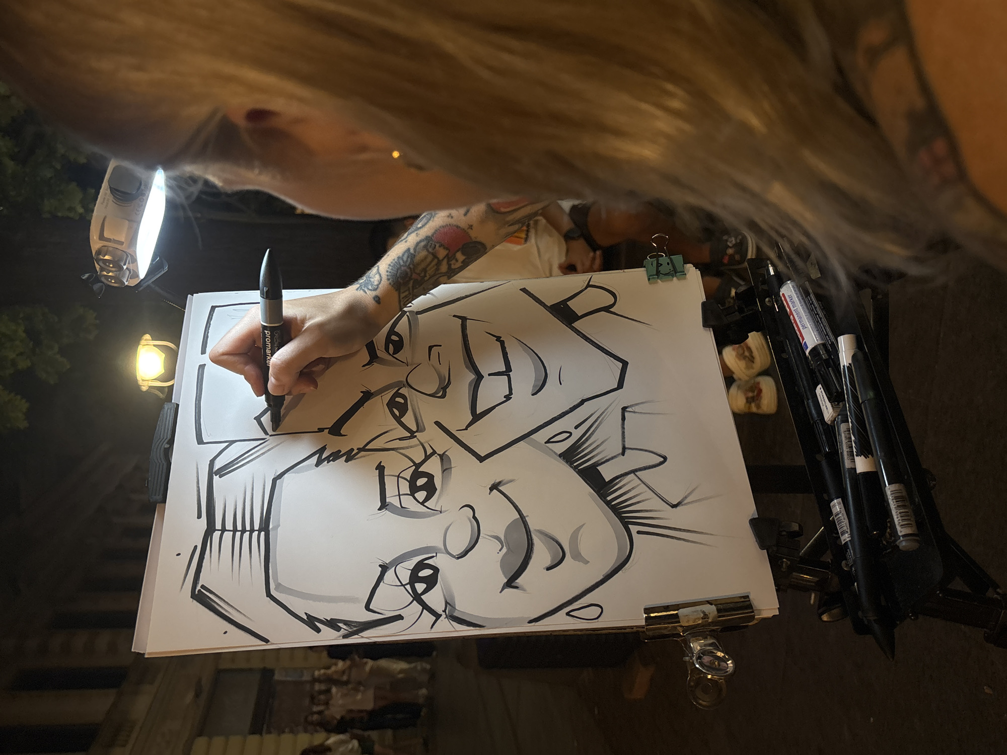 An artist with blonde hair and tattoos on her arm is drawing a caricature of two people on a whiteboard, which is propped up on an easel. The artist is holding a black marker in her right hand and is in the process of drawing the right eye of the person on the right. The whiteboard features a caricature of a man and a woman, both with exaggerated features. The man has a large smile and prominent teeth, while the woman has large eyes and full lips. The drawing is done in black ink with some shading.
Behind the easel, a group of people are standing and watching, suggesting an outdoor setting. The background shows the facade of a building and trees, with a street lamp illuminating the scene. The artist's workstation includes a tray filled with various markers and drawing tools. The artist appears to be a woman, and her hair is visible over her right shoulder. The overall impression is of a lively street art scene.
The artist is likely working on Knez Mihailova Street in Belgrade, Serbia, a popular pedestrian zone known for its street performers and artists. The architectural style of the building in the background and the general ambiance of the scene are consistent with this location.