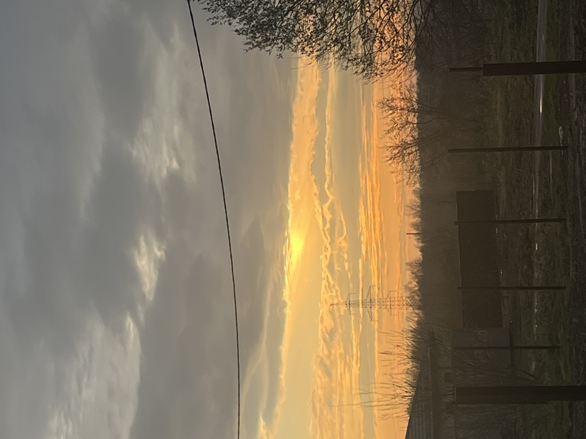 This image captures a dramatic sunset or sunrise over a rural landscape, likely in Раздолье, Russia. The sky dominates the frame, transitioning from dark, greyish clouds on the left to an intense, fiery orange and yellow glow on the right, indicating the low position of the sun. Streaks of illuminated clouds, possibly elongated contrails, span across the brighter sections of the sky.
A dark power line runs diagonally from the upper left toward the center-right. In the foreground and midground, bare tree branches are silhouetted against the sky, suggesting late autumn or early spring. The horizon line is visible across the lower portion of the image, revealing a hazy, dark landscape. An electricity pylon stands silhouetted against the bright sky, indicating human infrastructure within the natural setting. Below, the ground appears dark and indistinct, with further silhouetted trees and several dark, vertical structures resembling fence posts. A faint glint of light in the far distance on the right might indicate a road or water body.
The time of day is clearly either sunrise or sunset, characterized by the warm, intense colors and low sun angle. The weather appears partly cloudy. No discernible text is visible in the image. The scene evokes a sense of natural beauty and tranquility despite the presence of power infrastructure.