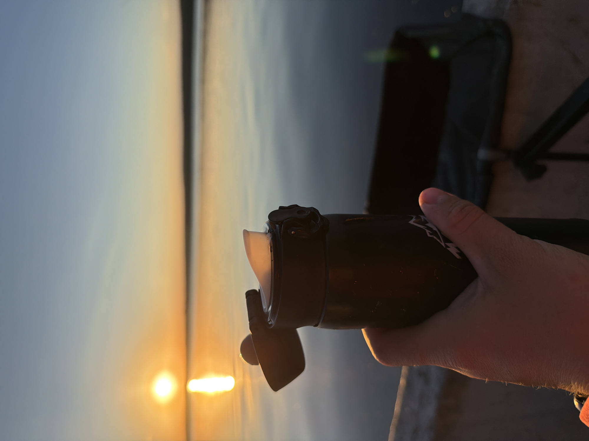 A person's hand holds a black thermos with an open lid against a backdrop of a sunset over a body of water. The sun, a bright orange orb, is low on the horizon, casting a warm glow on the water and the sky, which transitions from a vibrant orange to a soft blue. The water is calm, reflecting the colors of the sunset. In the background, out of focus, is a dark, rectangular object that appears to be a collapsible chair or bag. The scene suggests a peaceful moment outdoors, possibly at dusk. The location is identified as Chaikovsky, Russia.