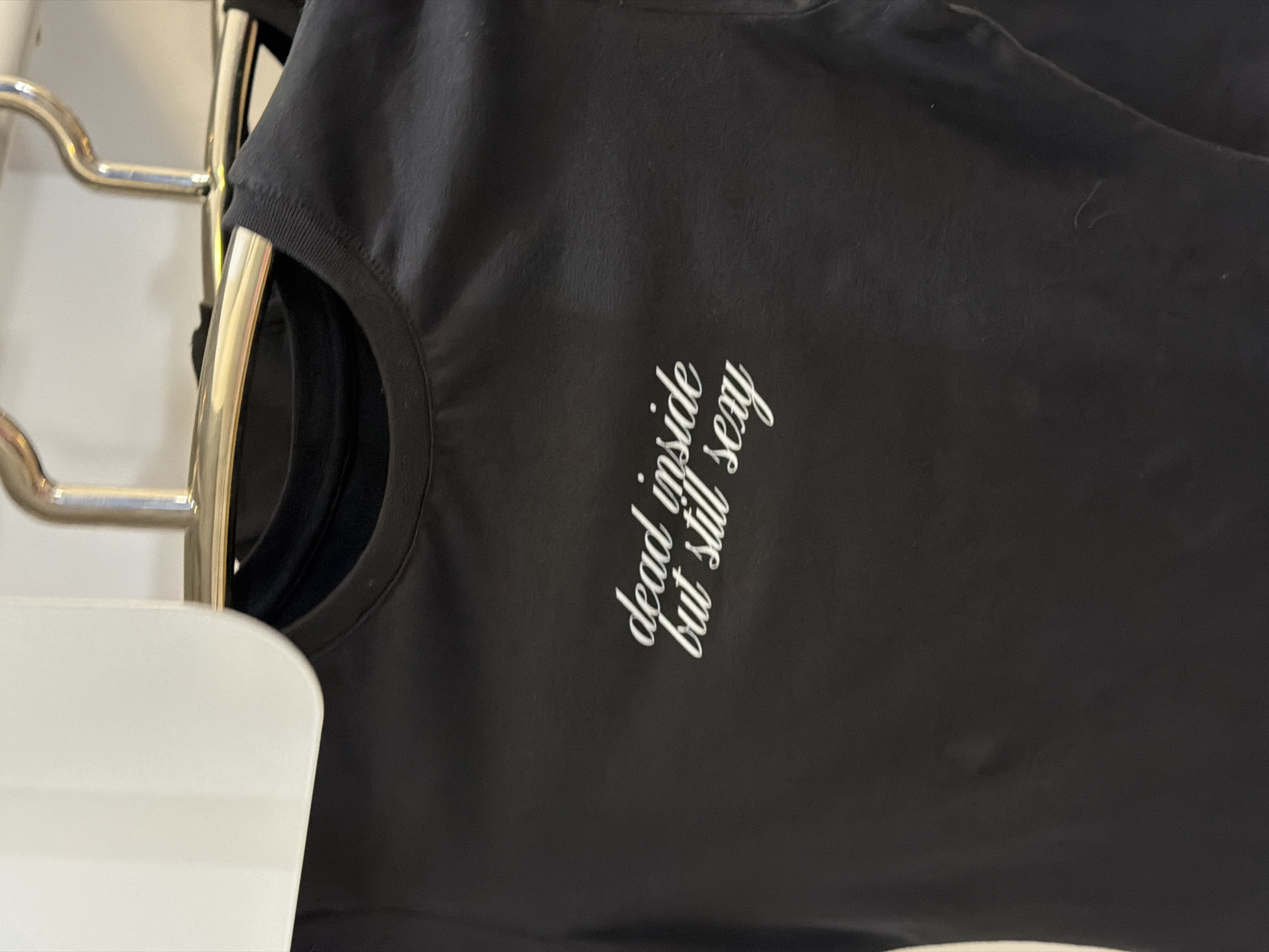 A black t-shirt hangs on a metal rack. The shirt has the white text "dead inside but still sexy" printed on it. The image is cropped, showing the top part of the shirt, and the metal rack is visible on the left side. A white object is partially visible in the lower-left corner of the image. The location or city is not discernible.