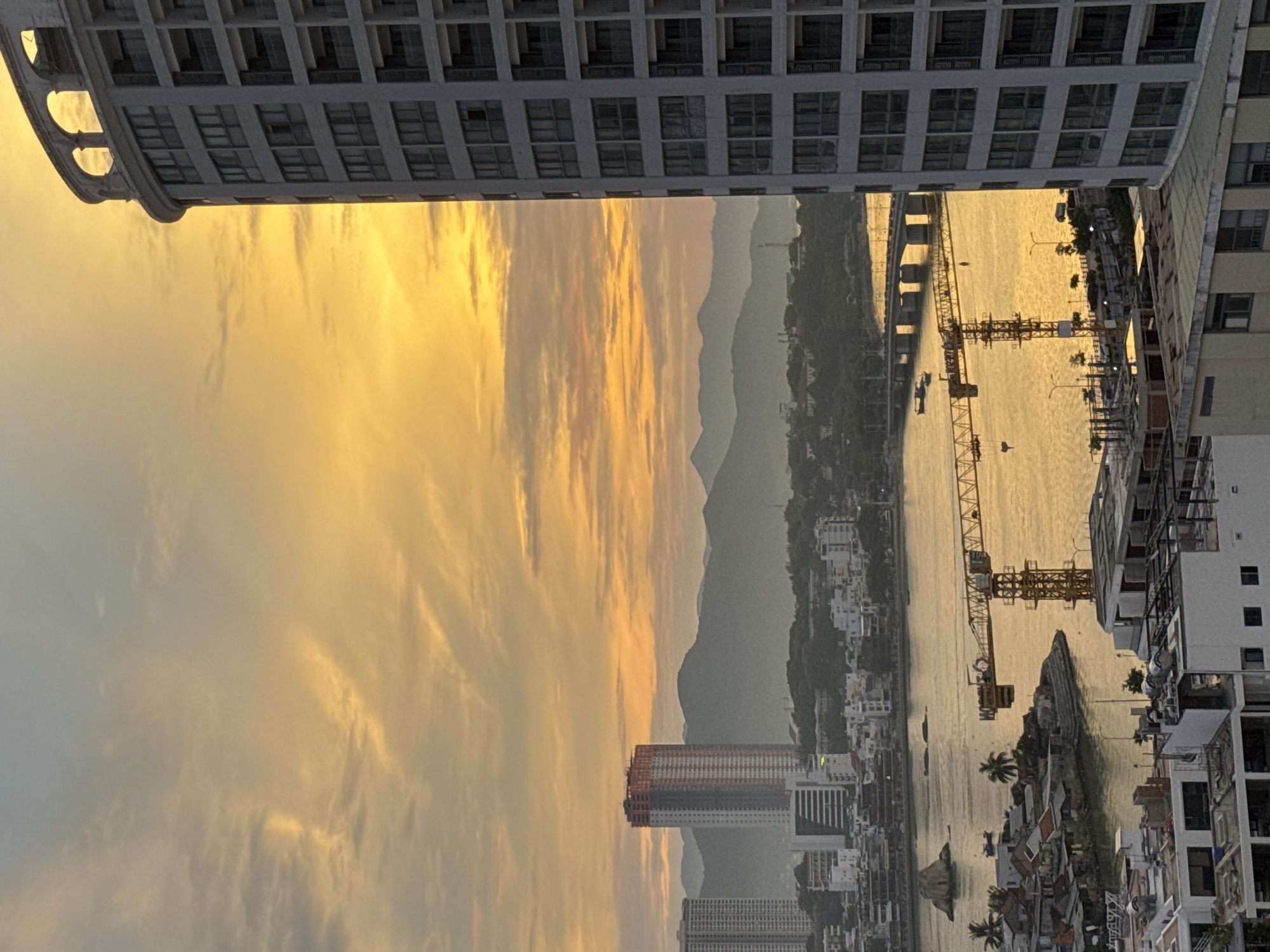 This is an eye-level, full shot of a city skyline in Nha Trang, Vietnam, at sunset. The sky is a vibrant orange and yellow, with soft clouds. In the foreground, a modern, white high-rise building with a curved facade dominates the right side of the frame. Its numerous windows are visible.
To the left of the high-rise, a body of water, likely the Cai River, reflects the warm tones of the sunset. A large yellow construction crane is positioned over the water, with its boom extending horizontally. Across the river, the city sprawls, featuring smaller buildings, trees, and a distant bridge. In the background, a range of hazy, green mountains forms a backdrop to the urban landscape. Another tall, orange and white building is visible further inland, amongst other city structures. The image conveys a sense of urban development against a natural, scenic backdrop.