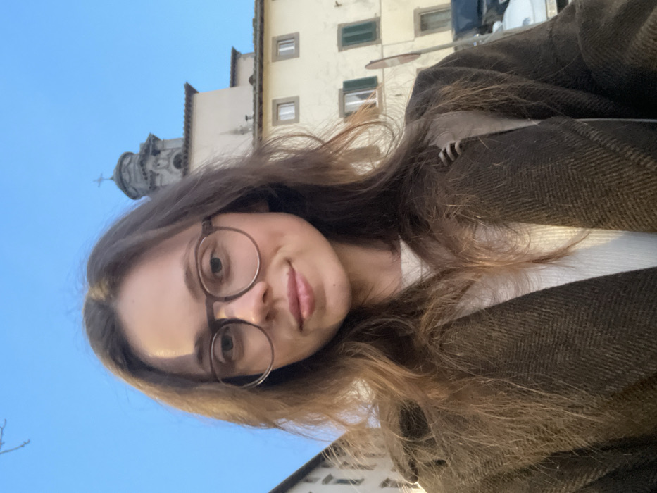 A young adult female is depicted in a close-up, chest-up selfie, looking directly at the camera with a slight smile. She has long, light brown hair and wears dark-rimmed round glasses, a light-colored ribbed top, and a dark brown, possibly herringbone-patterned, overcoat.
The background reveals an urban setting under a clear, light blue sky in Castel del Piano, Italy. To the left, a portion of a light-colored building is visible. In the mid-ground, a multi-story, light-colored building with a darker roofline is prominent. This building features several rectangular windows, with some on the lower visible floor having closed green shutters and upper-floor windows appearing open or un-shuttered. Above and behind this structure, a stone bell tower with a domed top surmounted by a cross is visible, indicating a church or similar religious edifice. A vertical pole with a circular sign (partially obscured, red border visible) stands in front of the building, and a dark-colored vehicle is partially visible behind the pole, to the right of the woman.