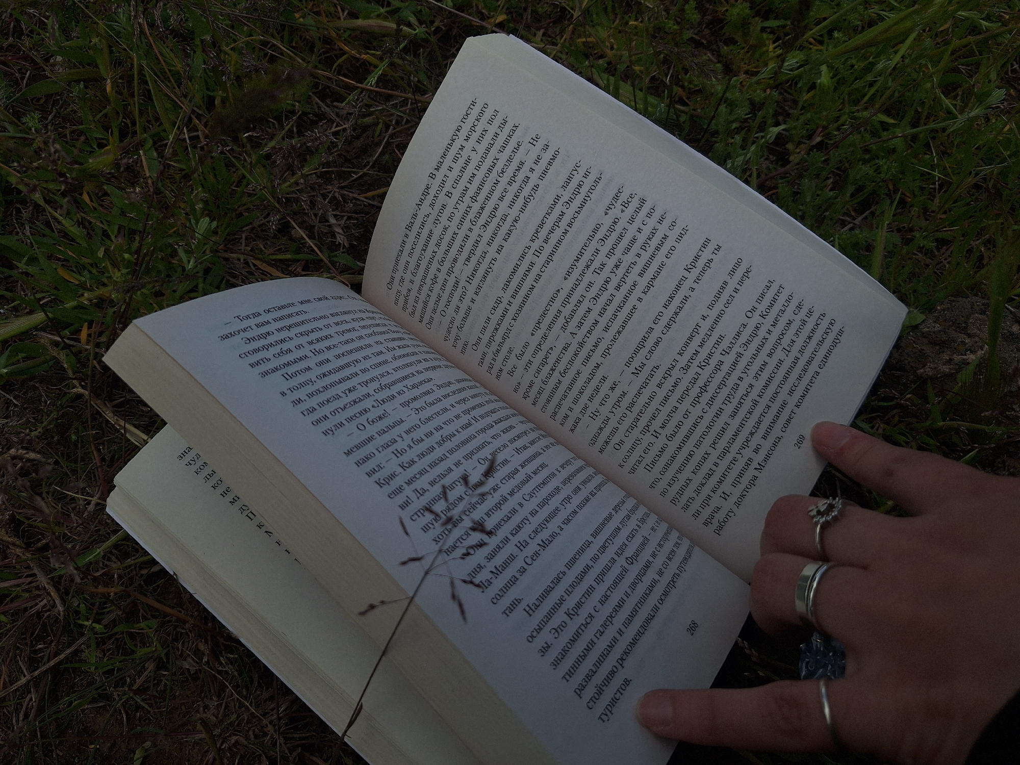A low-angle, medium shot captures an open book lying on a bed of grass, with a person's hand resting on the right-hand page. The book's pages are filled with text in what appears to be the Russian language. The visible page numbers are 268 and 269. The hand, adorned with multiple rings, holds the book open. The grass is dense and uneven. The lighting is subdued. There is no indication of the location being in the city of Tlik, Armenia.