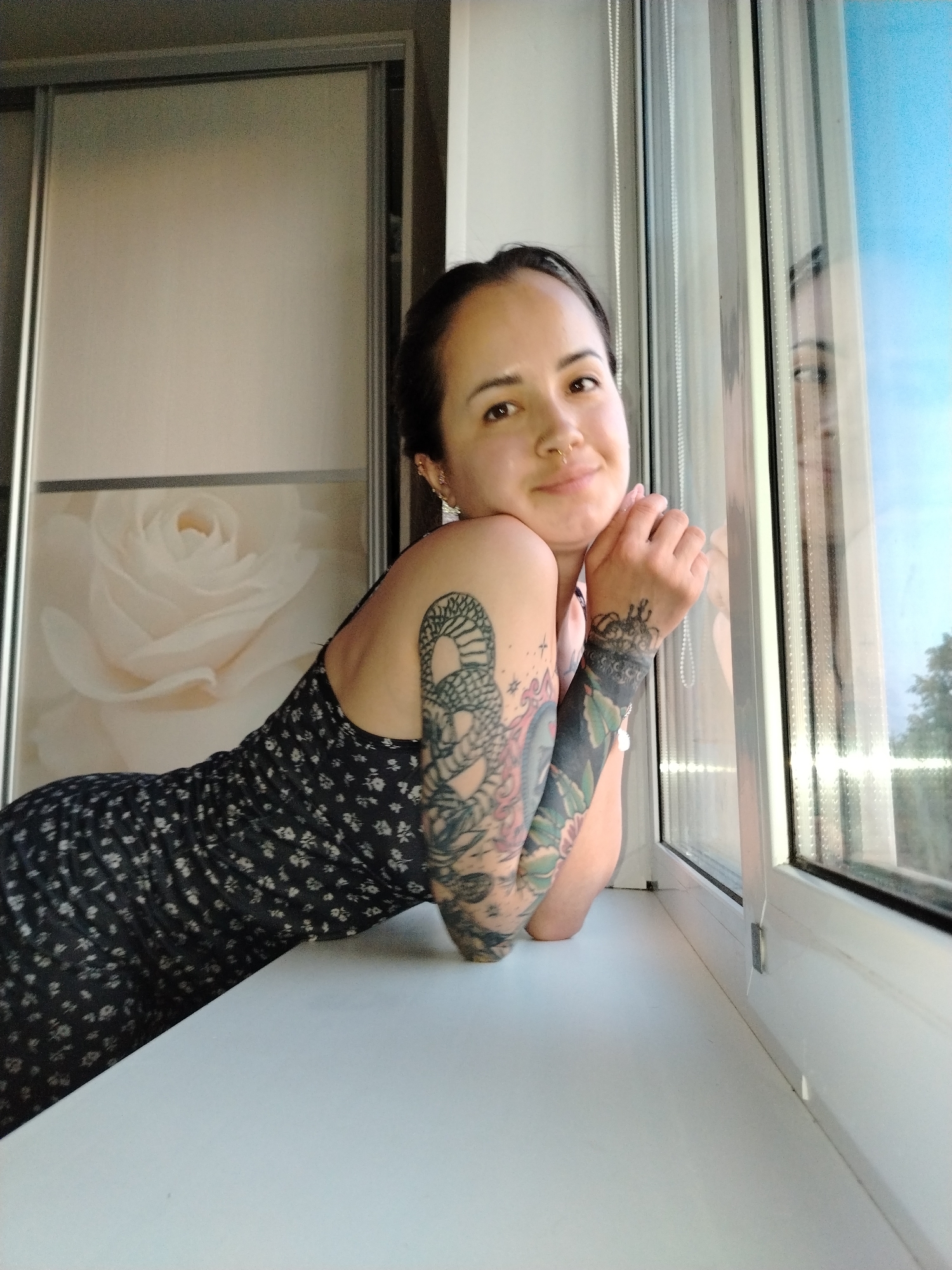 A woman with tattoos on her arms is leaning on a window sill in a room. She is wearing a dark floral dress. A closet with a floral design is visible behind her. The window looks out onto a sky with a hint of greenery, suggesting a residential area. Noviy, Russia is not discernable from the visual details.