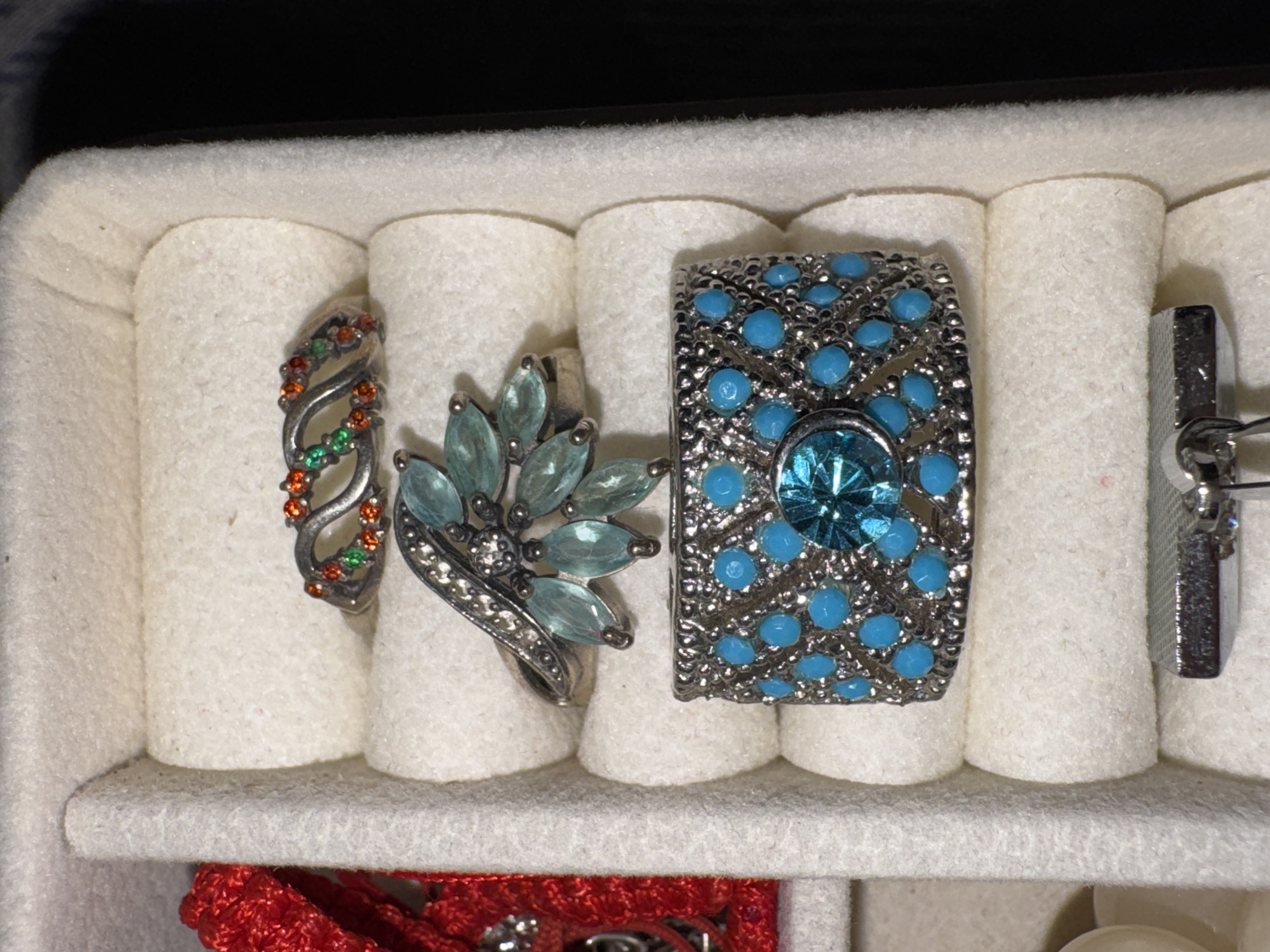 The image shows a top-down view of a jewelry box compartment containing several rings. In the top section of the compartment, a silver ring is visible, adorned with small orange and green gemstones arranged in a wave-like pattern. Below this, another silver ring is positioned, featuring a cluster of light blue, marquise-shaped gemstones set in a fan or feather design, with smaller clear stones around the center. The largest ring, located at the bottom of the compartment, is a wide silver band encrusted with numerous small, bright blue circular stones, and a prominent, faceted, bright blue gemstone in the center. A partially visible red string or ribbon is on the far left of the image, and another silver object, possibly a ring or jewelry clasp, is at the very bottom. The jewelry box compartment is lined with a soft, white material. There is no indication of a city in Яромінскі сельскі Савет, Belarus.