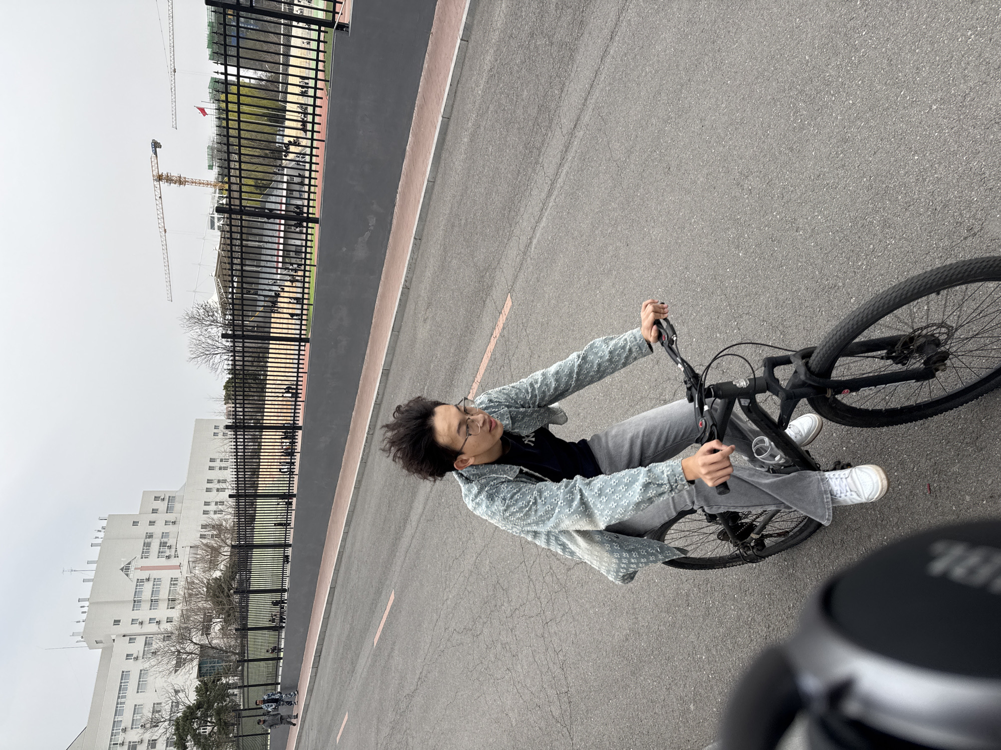A young adult wearing glasses, a light blue jacket, a black t-shirt, and gray pants is riding a black bicycle on an asphalt road. The bike rider is facing forward with a slight turn to the left. In the background, there's a tall white building, a black metal fence, and construction cranes. The location is Zhongtun, China.