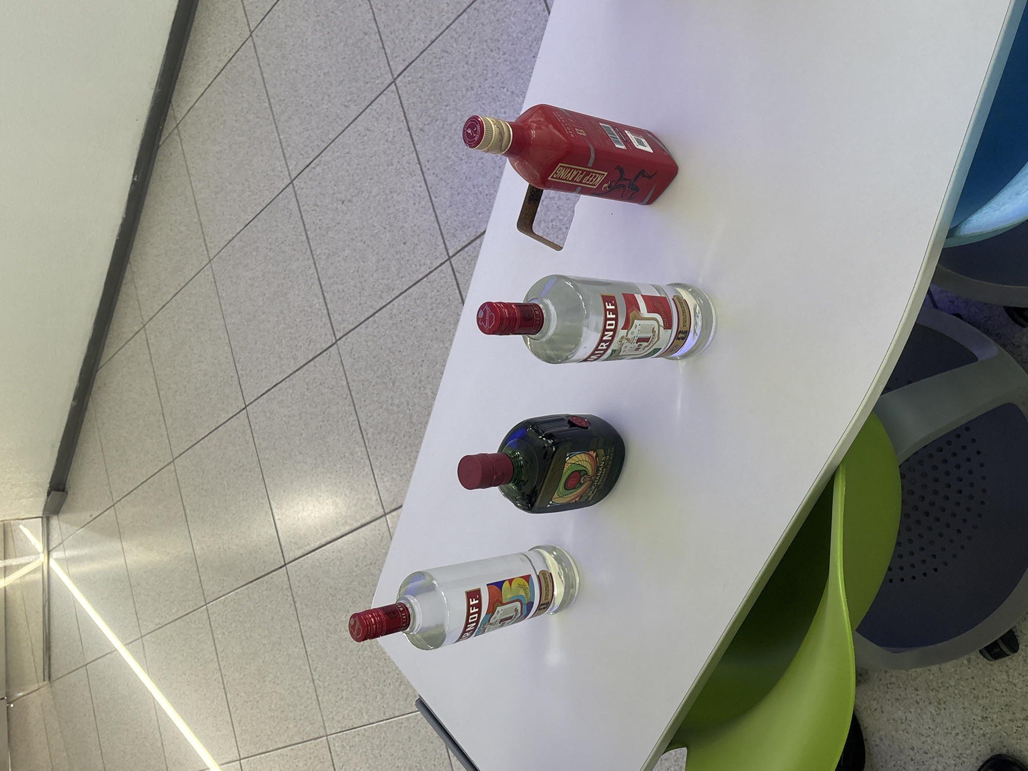 A high-angle shot shows four bottles of alcohol lying horizontally on a white table. A bottle of Smirnoff vodka, a bottle of Buchanan's scotch whisky, and a bottle of Keep Playing rum are visible. The location cannot be determined from the image.