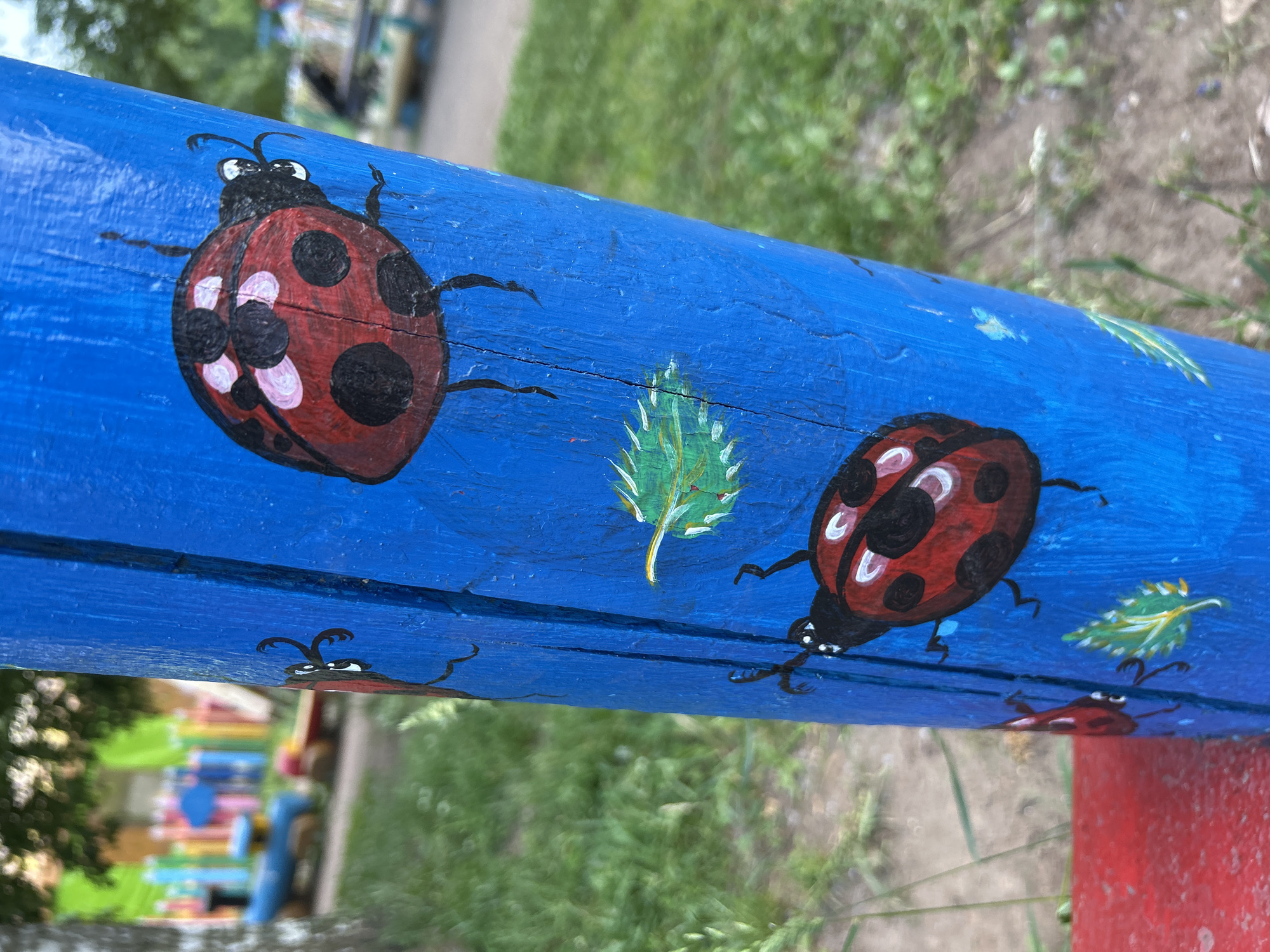 The image captures a close-up of a bright blue painted wooden pole, likely part of a playground structure in Chaikovsky, Russia. The pole is adorned with hand-painted ladybugs and green leaves. Two ladybugs are prominently featured; one near the top, facing right with black spots on its red shell, and another near the bottom, also facing right. Several small green leaves are painted around the ladybugs. The background is blurred, showing a grassy area and what appears to be a colorful playground with fences and structures. The focus is on the painted details of the pole.