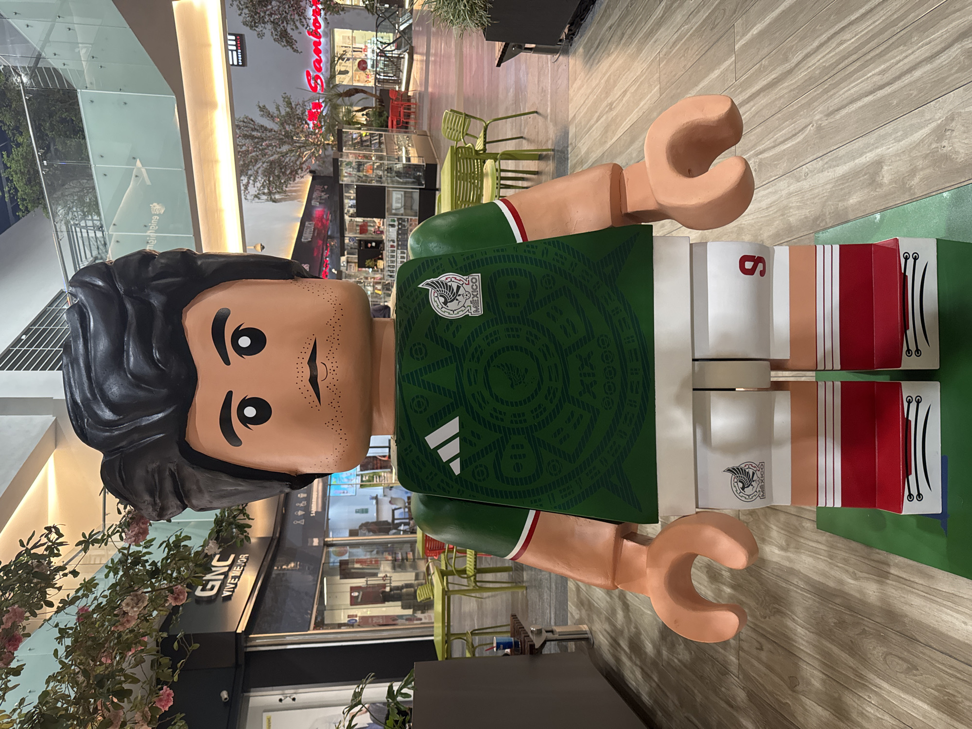 The image shows a large Lego figure of a male soccer player wearing a Mexico jersey. The jersey is green with the Mexican coat of arms on the front. The shorts are white with red stripes on the sides, and the number 6 is visible on one of the shorts. The Lego figure has black hair, brown skin, and black dot eyes. The figure is located in a shopping mall. The mall has multiple stores and seating areas. The location is Puebla, Mexico.