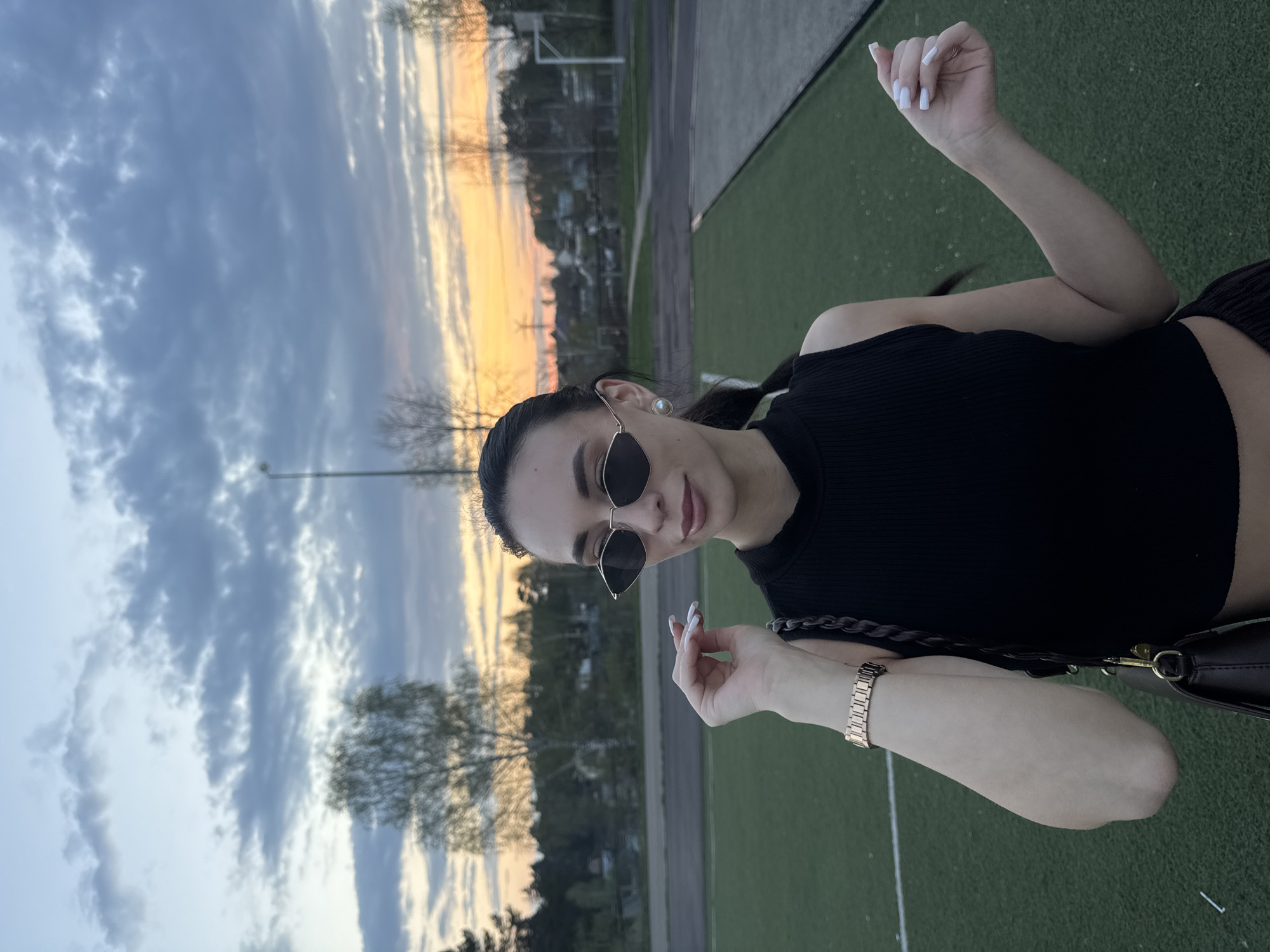 A woman with dark hair pulled back in a ponytail is posing outdoors at dusk in Chaikovsky, Russia. She is wearing a black sleeveless top, sunglasses, and a gold watch. She is positioned on a green surface that appears to be an artificial turf field. Behind her, the sky displays shades of blue, gray, and orange, suggesting a sunset or sunrise. There are trees and buildings visible in the background, along with a tall light pole and a soccer goal.