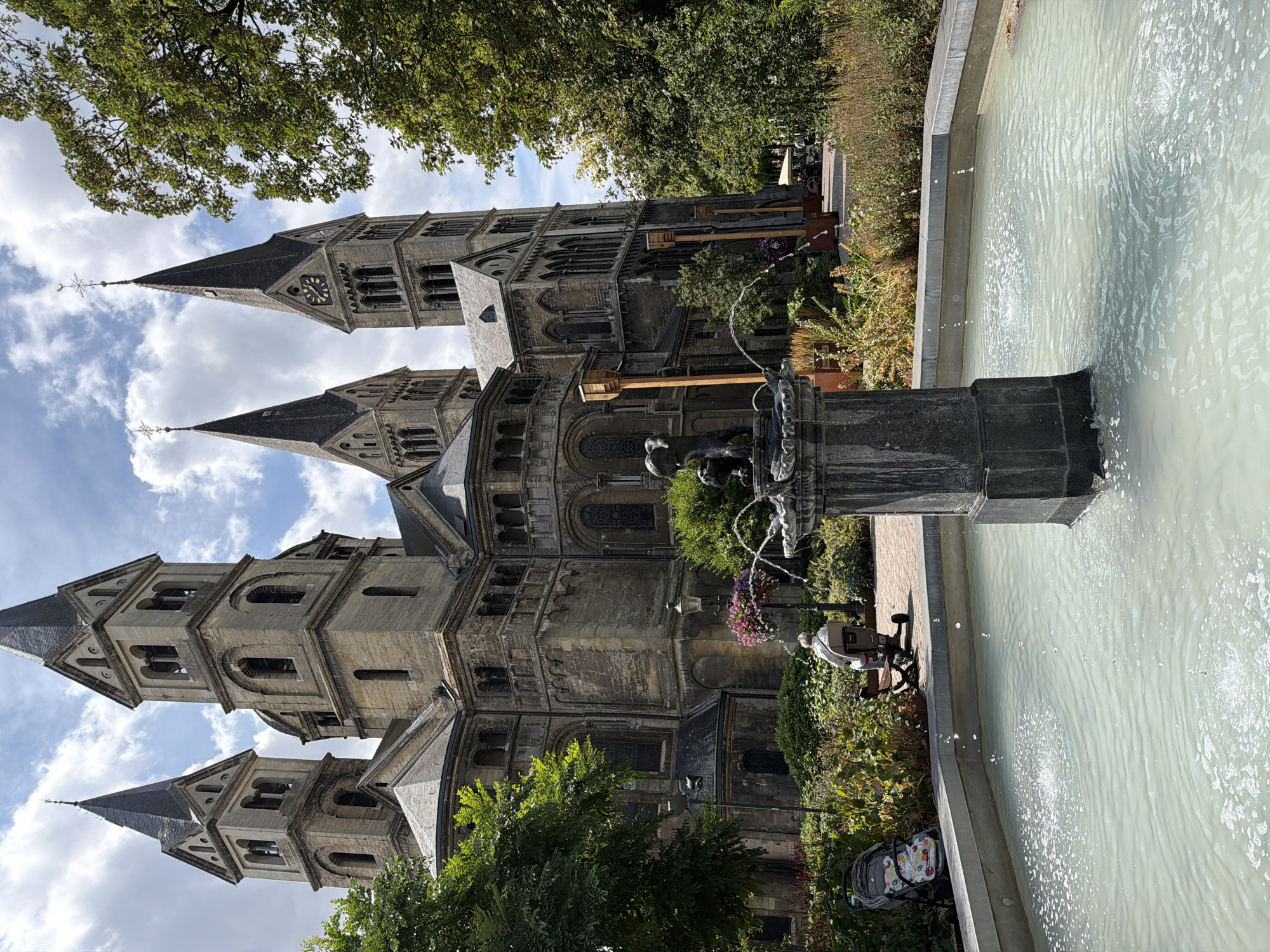 In Roermond, Netherlands, a light-colored fountain with spraying water occupies the foreground. Behind it, a person sits on a mobility scooter in a garden area with lush greenery and blooming flowers. A baby stroller is parked nearby. The main subject is a large, old stone church with multiple spires, Romanesque architectural features, and a clock on one of its towers. Trees frame the right side of the image, and a cloudy sky is visible overhead.