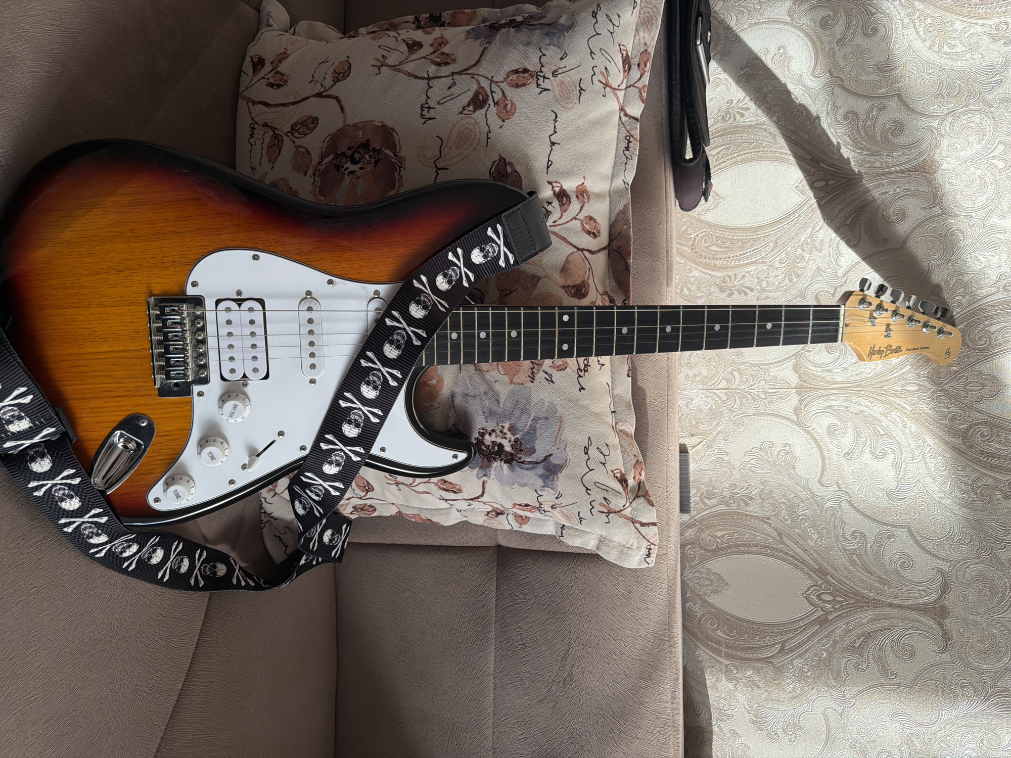 This media file features an electric guitar, likely a Stratocaster-style model, resting on a plush sofa. The guitar has a sunburst finish with shades of brown, orange, and yellow. It is equipped with a black guitar strap adorned with a pattern of white skulls and crossbones. The guitar's white pickguard holds three single-coil pickups and several control knobs and a selector switch. The neck of the guitar is visible, with frets and dot inlays. The headstock displays a logo that reads "Harley Benton Standard Series."
The guitar is positioned diagonally across the frame, with its body resting on a patterned cushion. The cushion has a floral design in muted tones of brown, beige, and grey, with some script-like markings intermingled. Behind the cushion and to the right of the guitar, a textured wallpaper with an ornate damask pattern is visible. The scene appears to be indoors, illuminated by natural light casting shadows from the guitar and strap onto the wall. The soft furnishings and the presence of a guitar suggest a casual, domestic setting. There are no people visible in the media file. The overall mood is one of stillness and perhaps relaxation or anticipation, given the instrument's presence. The time of day is likely daytime due to the bright lighting and distinct shadows. The location context provided is Baku City, Azerbaijan.
