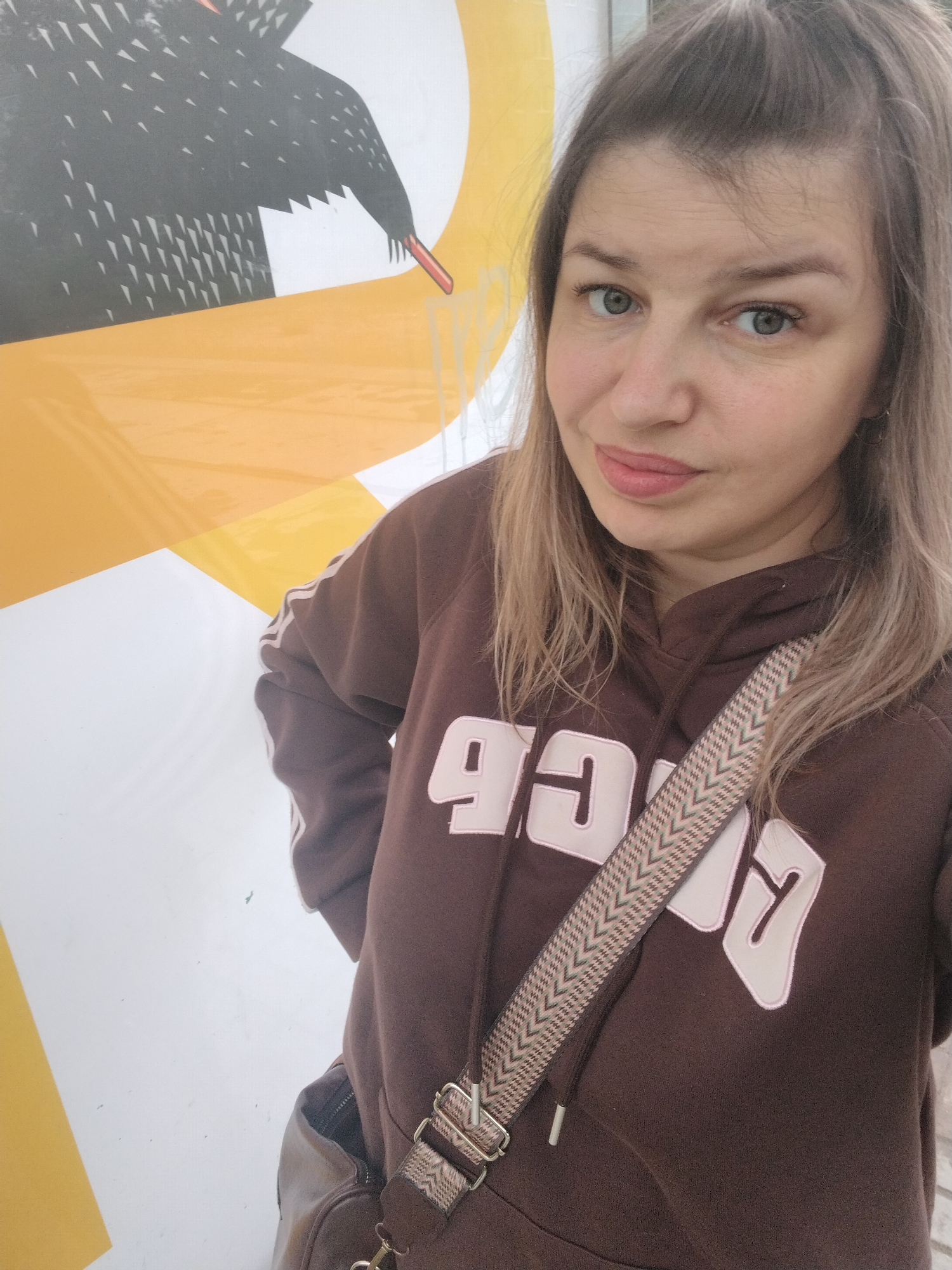 In Yaroslavl, Russia, a woman with blonde hair pulled up in a high ponytail and light green eyes stands in front of a large graphic display. She is wearing a brown hoodie with white lettering and a patterned strap from a dark brown bag crosses her chest. The graphic behind her features a large black bird with sharp white markings and a yellow and white background. The overall scene is bright and appears to be outdoors.