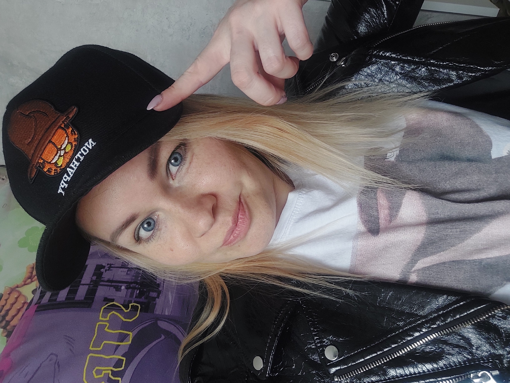 A woman with light blonde hair and blue eyes poses for a selfie. She is wearing a white t-shirt underneath a black, patent leather jacket. She is also wearing a black baseball cap with an orange cartoon character embroidered on it. Her left hand is raised, with her index finger pointing at the brim of the cap. The background includes a patterned surface, possibly a pillow or other soft furnishing, with shades of purple, pink, and white. The photo appears to have been taken indoors. There is no information to support the image being taken in Chaikovsky, Russia.