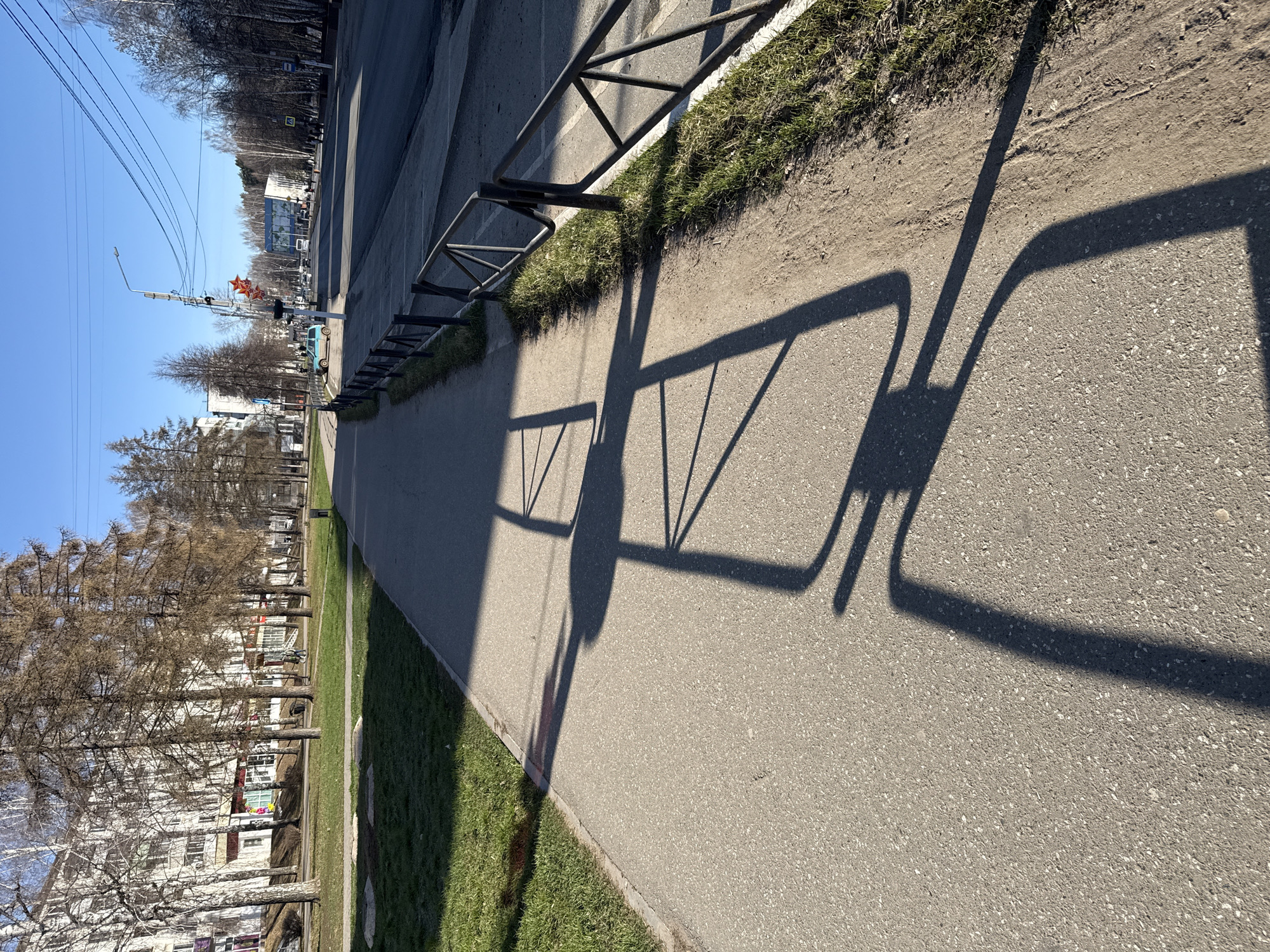 A street scene during daylight in an urban environment. The camera is oriented along a sidewalk adjacent to a road. A black metal barrier runs between the sidewalk and a narrow strip of grass. Shadows of the barrier are cast prominently on the sidewalk. Beyond the road are buildings and trees in the background. The sky is blue and mostly clear. A traffic light is visible in the distance. A teal colored automobile is also in the background.