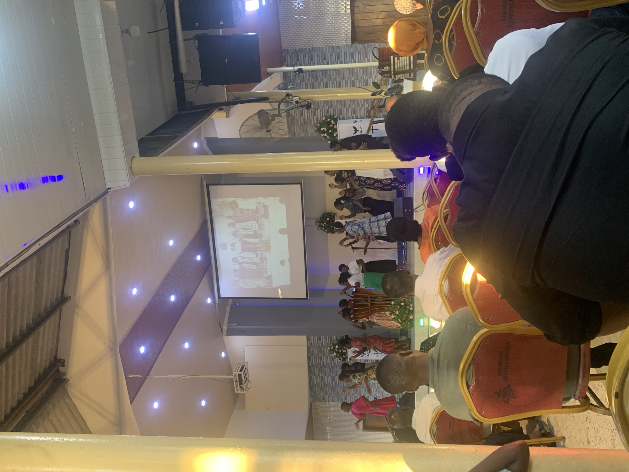 The image shows a gathering of people inside a building with a stage and a projection screen. The location is Dar es-Salaam, Tanzania. People are seated on red chairs facing a stage. A projection screen displays images on the stage. There are several pillars supporting the roof. The lighting is provided by ceiling lights. People are engaged in an event or presentation.