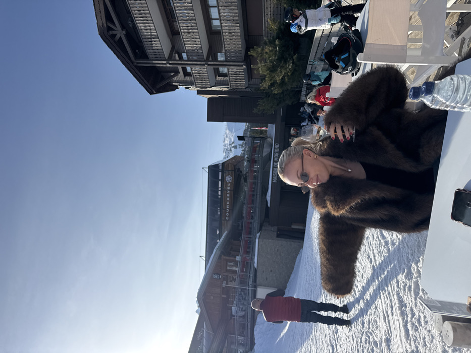 A sunny, snow-covered outdoor scene at a ski resort in Courchevel, France, is depicted. A blonde woman, with hair pulled back, wearing sunglasses and a brown fur coat, is seated at a white outdoor table, holding a stemmed glass containing a light-colored beverage in her right hand. Her nails are painted red. On the table, to her left, are two cylindrical shakers and a dark smartphone. To her right, a black ski helmet and a plastic water bottle with two clear bottles are visible. Behind her, a male individual in a red vest and dark clothing walks uphill on a snow-covered slope. In the midground, other patrons are seated at outdoor tables. Visible buildings include a multi-story wooden structure with balconies on the right, and a building in the center labeled "Maison de Moriond." Further left, a wooden structure is visible beneath a ski lift labeled "COURCHEVEL" with a snowflake emblem. Snow-covered mountains and a clear blue sky complete the background.