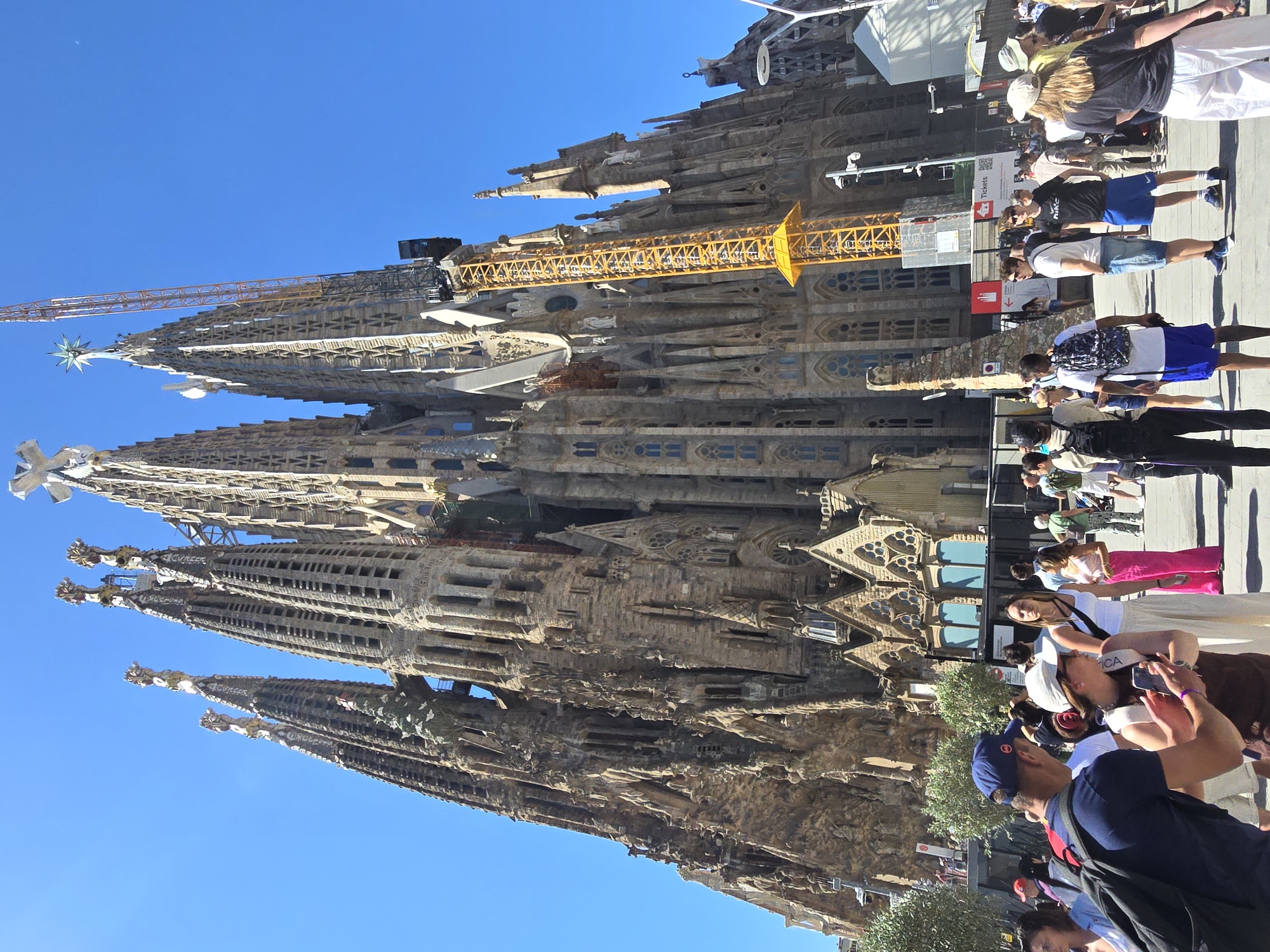 The image depicts a low-angle, full-shot view of the Sagrada Familia, a Roman Catholic minor basilica in Barcelona, Spain, on a clear, sunny day. The iconic structure, characterized by its towering spires and intricate facade, is partially obscured by a bright yellow construction crane positioned to its right. The foreground features a crowd of tourists gathered in an open plaza, some interacting with their phones or conversing. The people are dressed in casual summer attire. The architectural style of the Sagrada Familia is a blend of Gothic and Art Nouveau, with its stone facade appearing weathered and detailed. The sky is a vibrant, cloudless blue.