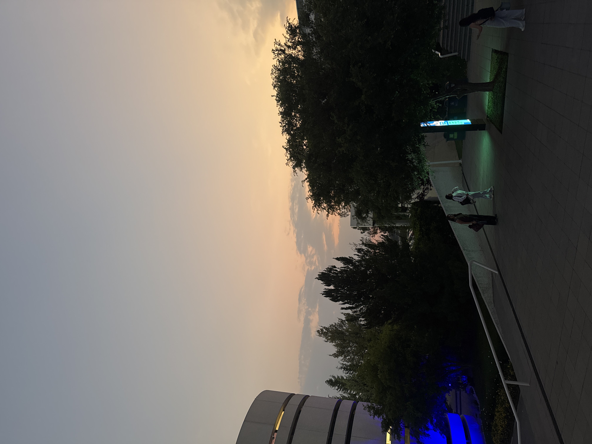 The image depicts an outdoor scene in what appears to be an urban environment at dusk or twilight. The sky transitions from a pale gray to an orange-tinted hue, indicating the setting or rising sun. Trees are scattered throughout, silhouetted against the colorful sky. On the right, there is a paved walkway with a few people walking. Blue lights illuminate a structure, possibly part of a building or art installation, and some greenery along the walkway. A curved building is visible in the bottom of the frame. The location is Puebla, Mexico.