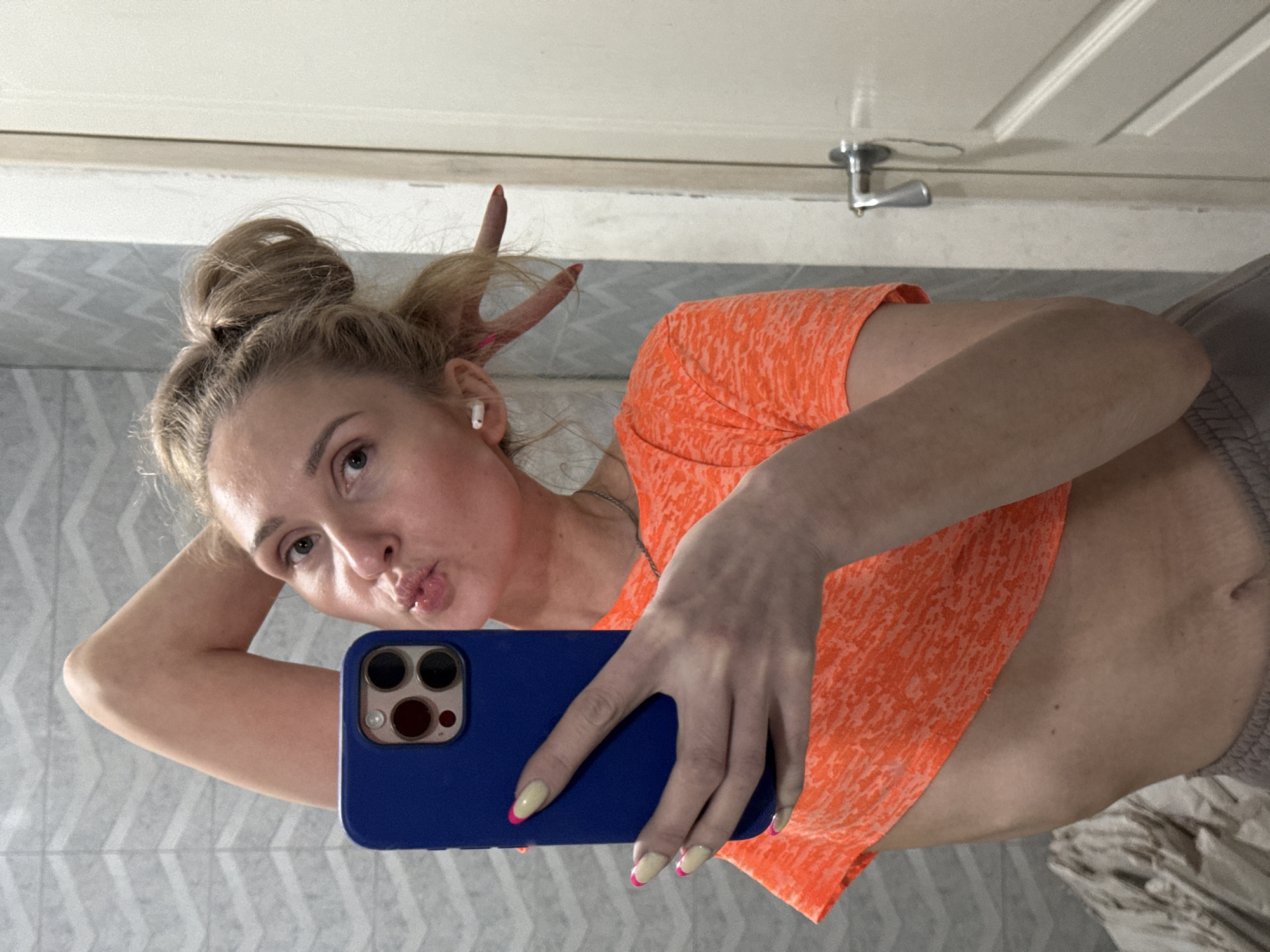 The image depicts a woman with blonde hair styled in a high bun, seemingly taking a selfie in a bathroom. She is wearing an orange t-shirt and is holding a blue smartphone. Her nails are painted with a light color and a pink french tip. The background shows a tiled wall and a door with a silver handle. She is making a kissing face and has her fingers styled as antlers behind her head. There is no information in the image that allows the location to be pinpointed to Chaikovsky, Russia.