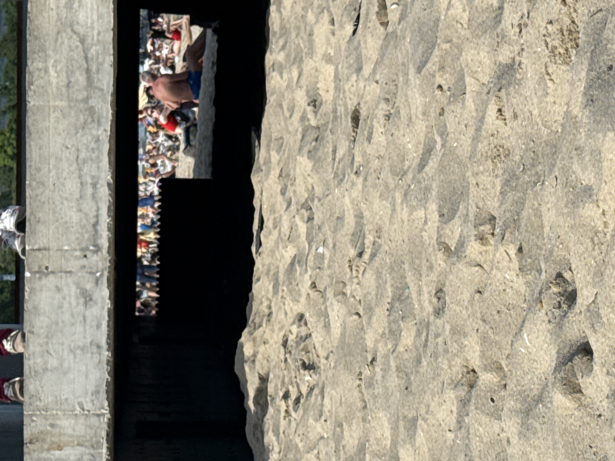 The image presents a composite view of a beach environment intersected by a concrete structure. In the immediate foreground, light-colored sand covers the ground, showing numerous footprints, small depressions, and scattered white shell fragments. Above this, a large, rough-textured grey concrete beam or slab spans horizontally, forming an overhead structure. Below this concrete, a dark, shadowy void exists, through which a rectangular opening reveals a sunlit beach scene.
Within this opening, numerous people are visible, primarily seated or reclining on the sand. A prominent figure on the right is a shirtless man with a tanned back, wearing blue shorts, seated on the sand. To his left, a woman in a red top and sunglasses is also seated. Other individuals are distributed across the visible beach area, engaged in passive activities like sunbathing. Faintly visible in the far distance through the opening are further beachgoers and possibly some beach structures.
Above the concrete structure, only the lower legs and feet of at least two individuals are discernible. On the left, feet clad in brown shoes with maroon accents are visible. On the right, feet wearing white athletic shoes are present. These individuals appear to be situated on top of the concrete structure. In the background above the structure, green foliage suggests a natural landscape beyond. The scene indicates a bright, sunny day. The location is an unknown city, unknown.
