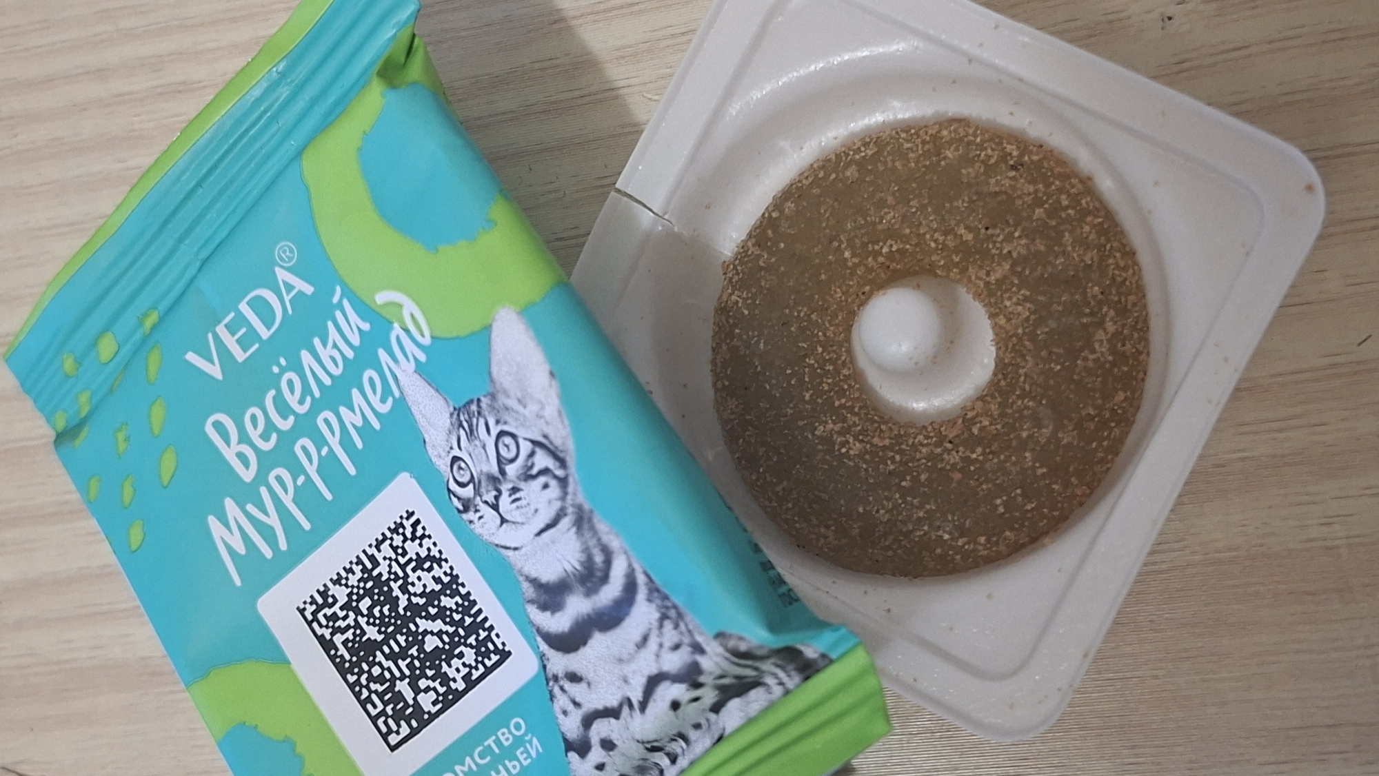 A close-up, overhead shot shows a pet treat shaped like a ring, covered in a brown crumbly coating, placed in a white plastic tray. To the left of the treat, a partially opened package of cat treats is visible. The package is primarily turquoise and green, featuring a QR code on the left side, and the text "VEDA Весёлый Мур-р-рмелад" in white lettering. An illustration of a cat's head is also present on the package. The items are positioned on a light brown, wooden surface, likely a table. The scene is lit evenly, suggesting indoor lighting. No people are present.