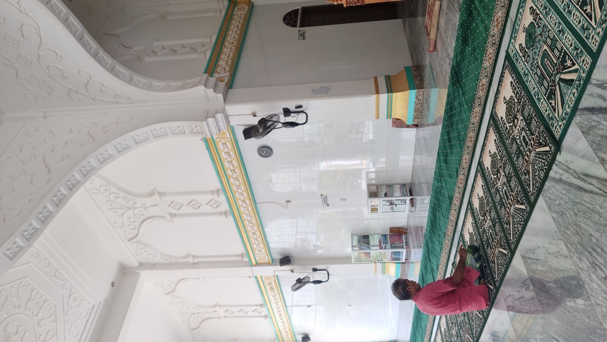 An overhead shot shows a man sitting on a prayer mat in a mosque. He is wearing a pink shirt and plaid pants. The prayer mat is green with a geometric pattern. The mosque has white tiled walls and a marble floor. There are arches and decorative moldings on the walls and ceiling. Two white shelves with books are visible in the background. Two fans are mounted on the walls. The man is facing to the left, and his hands are resting on his lap. The scene is brightly lit, suggesting it is daytime. The overall impression is one of peace and serenity. This mosque is located in the city of Lapang Timu, Indonesia.