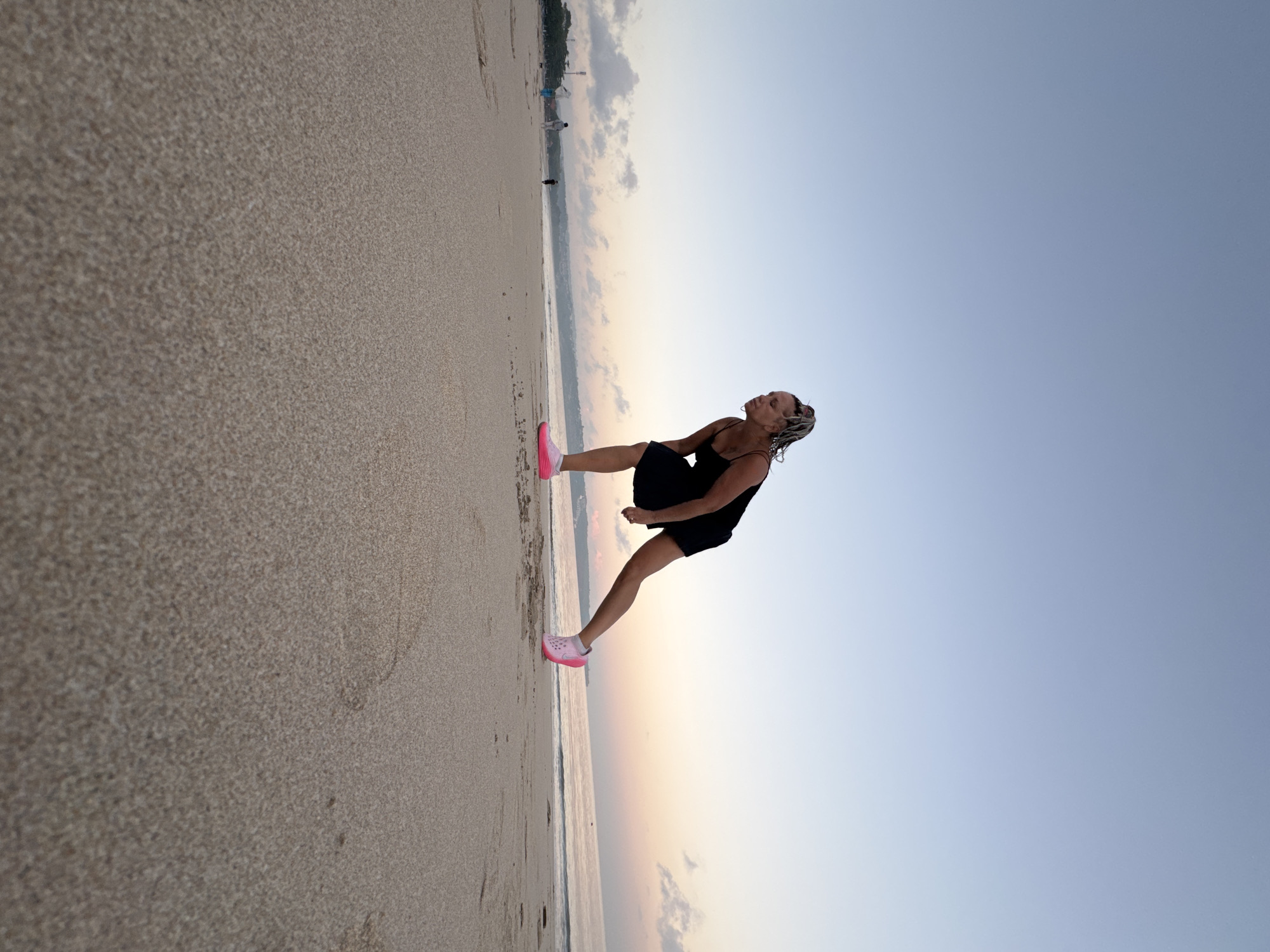 A woman with braided hair is stretching on a sandy beach in Kuta, Indonesia, as the sun begins to set. She is wearing a black tank dress and bright pink athletic shoes. The sky is a gradient of blue and soft orange, with a few wispy clouds near the horizon. The ocean is calm, with gentle waves lapping at the shore. In the background, other people can be seen walking along the beach. The sand in the foreground is detailed and textured, showing the fine grains and slight unevenness of the beach surface.