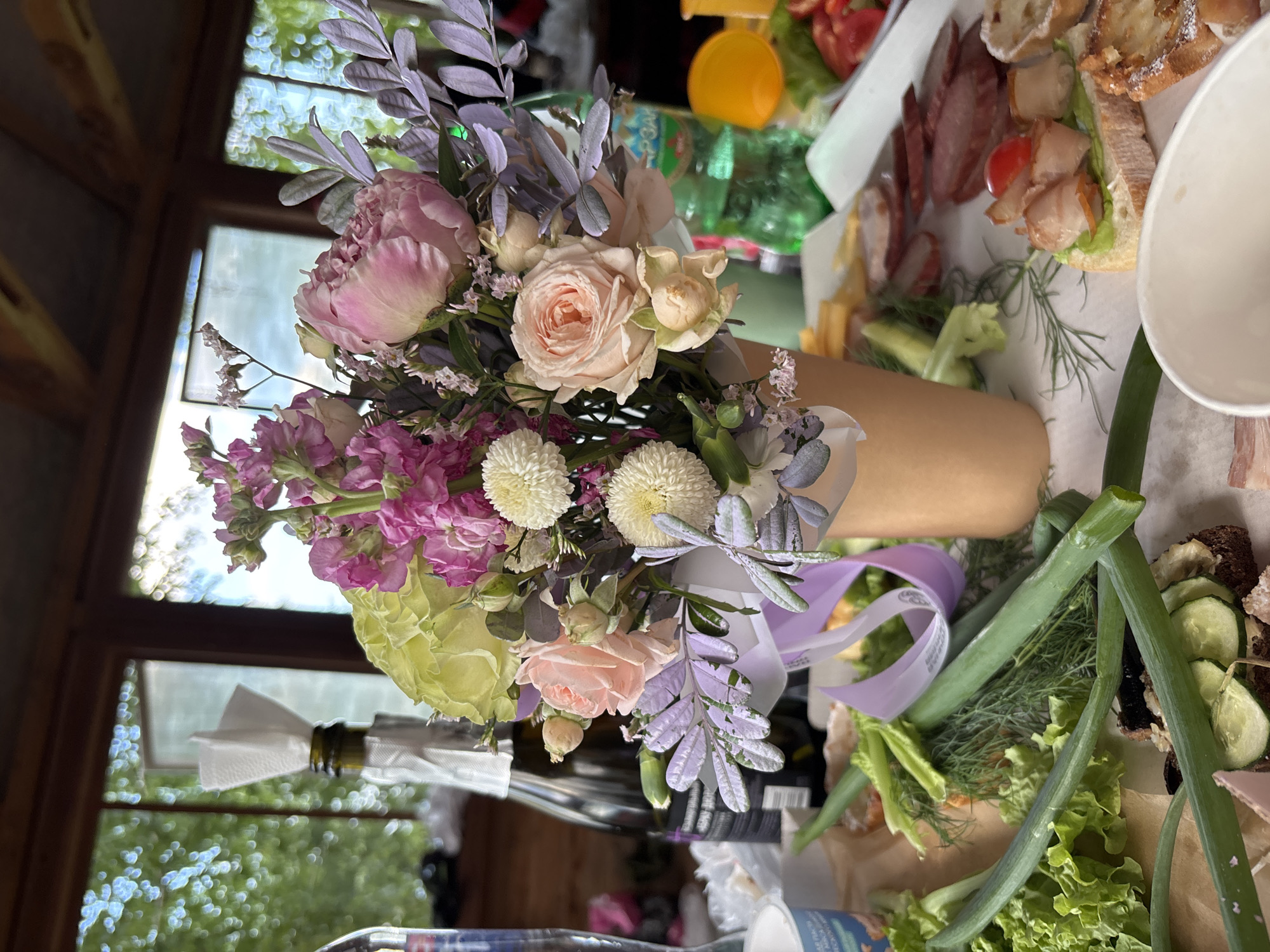 The image depicts an outdoor gathering or picnic, likely in Chaikovsky, Russia, given the context. A bouquet of flowers, featuring pink roses, white chrysanthemums, and purple foliage, is prominently displayed in the foreground, placed in a brown vase or holder. Beside the flowers, a bottle of champagne is visible, adorned with a white napkin.
The scene is set on a table laden with food, suggesting a celebratory meal or social occasion. Various food items are arranged, including sliced meats, small sandwiches with meat and vegetables, cherry tomatoes, sliced cucumbers, and green lettuce. Fresh herbs like dill and green onions are also present, adding to the appetizing display. In the background, a window with a view of lush green trees and bright sky indicates it's daytime with pleasant weather. The overall atmosphere appears relaxed and cheerful, hinting at a pleasant gathering among friends or family. Some visible text on a green plastic bottle in the background is illegible.