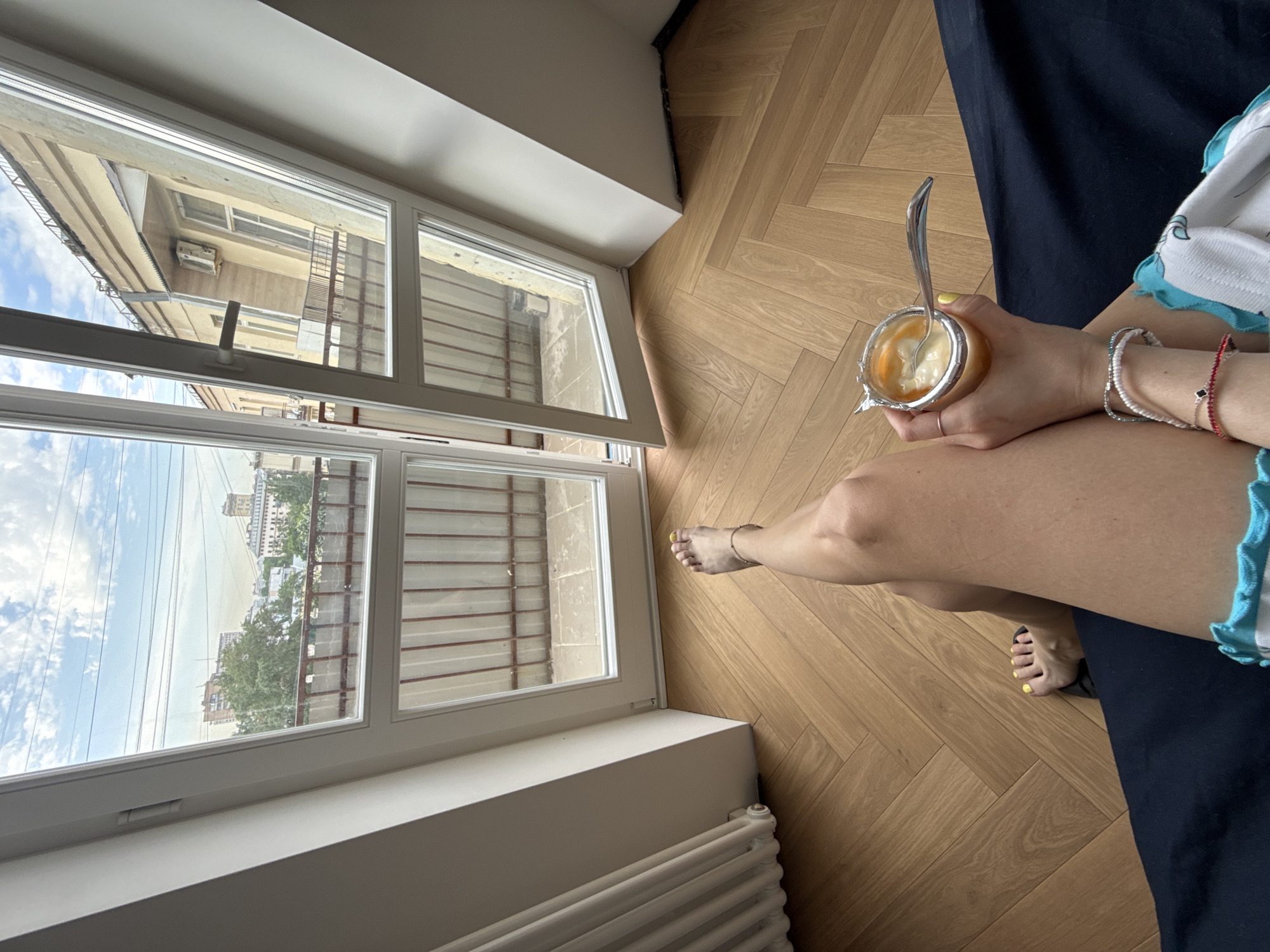A high-angle shot shows a person seated on a dark blue sofa or bed, with the lower portion of their body and a portion of their legs visible. The person is holding an open container of food with a spoon inside. Bracelets adorn their wrist. Outside the open window is a view of Moscow, Russia. Buildings and trees are visible through the open window, indicating a sunny day. The room has wooden floors laid in a herringbone pattern and a white radiator is visible near the bottom of the frame.