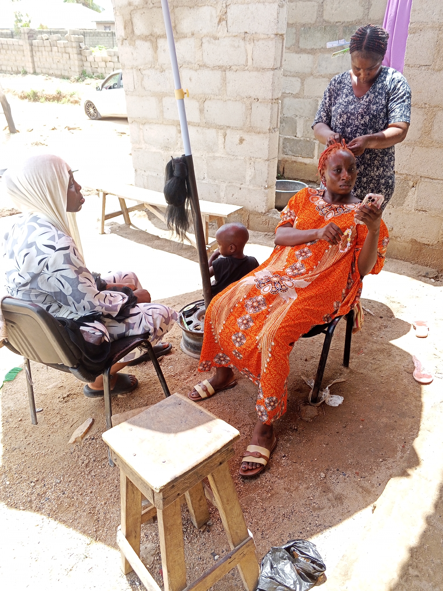 The image depicts a scene in an outdoor setting with two women and a child. One woman, wearing an orange patterned dress, is seated and appears to be using a mobile phone while another woman, wearing a dark patterned dress, stands behind her and braids her hair. A child in a dark shirt is sitting on the ground near the woman in the orange dress. Another woman, wearing a light-colored dress and headscarf, is seated in a chair to the left. A silver umbrella pole is positioned centrally in the scene. The background includes a partially visible building with brick walls. The ground is sandy. A wooden stool is in the foreground. The location is in the city of Unknown, Unknown.