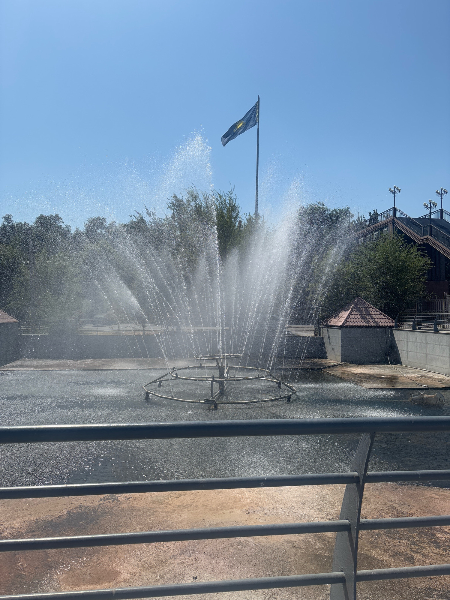 The image depicts a park fountain in Shymkent, Kazakhstan, on a clear, sunny day. The fountain is the central element, with water spraying outwards in a large, circular pattern. The water is captured mid-air, creating a shimmering effect against the blue sky. In the background, a flag with a dark blue field and a yellow sun emblem is flying from a tall pole. Trees line the background, and a building with a distinctive brown roof and decorative lamps is visible to the right. A metal railing runs across the foreground, obscuring a portion of the fountain and the ground. The overall scene suggests a public outdoor space, likely a park or recreational area within the city.