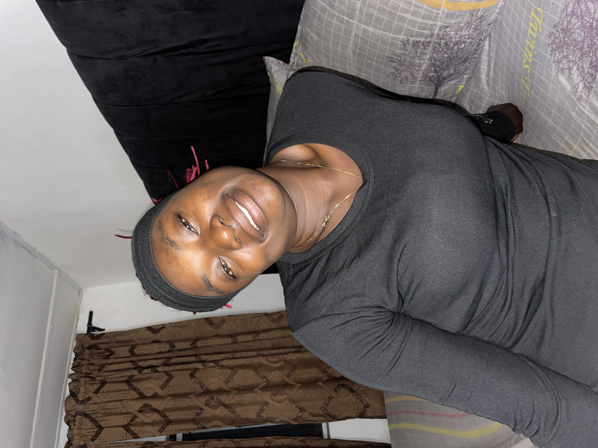 An indoor, eye-level portrait shows a young person with dark skin smiling and looking directly at the camera, their head tilted slightly to the left. This scene is located in a bedroom in Jos, Nigeria. The individual wears a black long-sleeved, ribbed top and a black headband, with reddish-pink strands of hair or decoration visible near the right temple. A thin gold chain necklace is worn around the neck. The person is seated or reclining on a bed, supported by pillows. To the left, a striped pillow displays horizontal bands of yellow, pink, and grey. To the right and beneath the person, a grey pillow and bed covering are visible, featuring a grid pattern and a stylized purple tree motif; partial text on the pillow appears to read "Twins Life". In the background, brown curtains with a repeating geometric pattern are drawn on the left, partially covering a window. A black curtain rod runs above them. On the right, a dark, likely black, padded headboard with vertical channeling is present. White walls are visible above the background elements, with a white ceiling and trim discernible in the upper left corner.