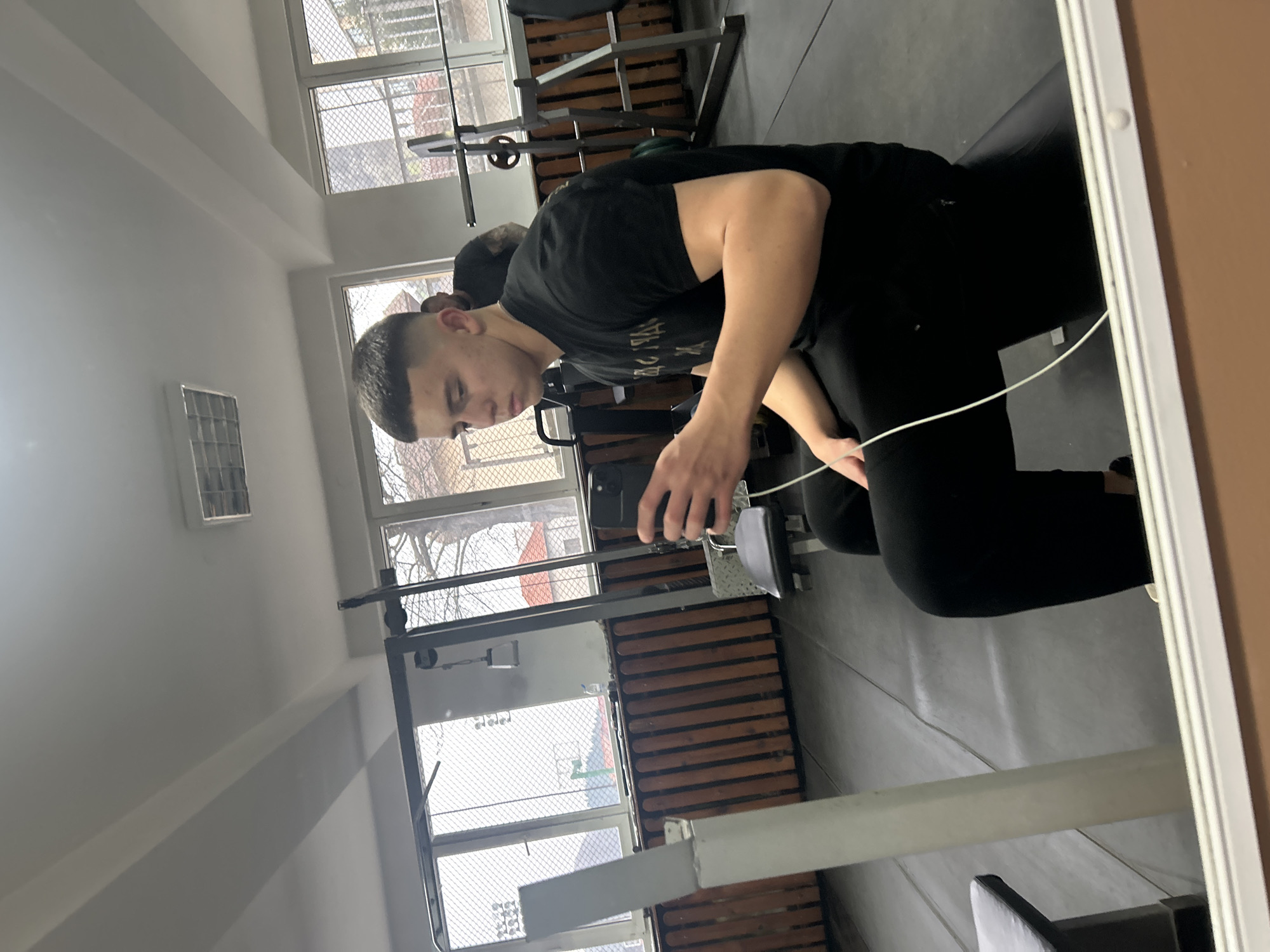 A young man with short, dark hair is indoors, seated on a weight training machine. He is wearing a black t-shirt and black pants. He is holding a black smartphone in his right hand and appears to be looking at it.
The setting seems to be a gym or workout area. Other weight training equipment is visible in the background. Large windows are present, offering a view of a tree and buildings.
The location is Zlatograd, Bulgaria.
