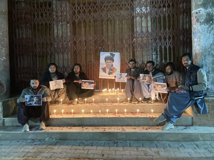 A nighttime scene in Quetta City Tehsil, Pakistan, depicts eight adult males participating in a solemn vigil on concrete steps outside a building. Seven individuals are seated or squatting on the steps, each holding a framed or unframed photographic portrait of a different male. One male on the far right is seated casually without holding a photo. All individuals are dressed in civilian attire, including traditional Pakistani clothing.
The steps are illuminated by approximately 25-30 lit white pillar candles, interspersed with scattered red rose petals. A large poster featuring a portrait of a man wearing a traditional cap and a patterned scarf is centrally displayed on the second step from the top. Text below the large portrait is partially visible, including "National Academic Movement Baloch". The background consists of a building facade, specifically a closed entrance or storefront secured by a dark, metallic, diamond-patterned roller shutter. The building's exterior appears to be a light-colored, possibly stucco or concrete, wall. The overall lighting is dim, primarily from the candles and ambient street sources, creating a somber atmosphere.