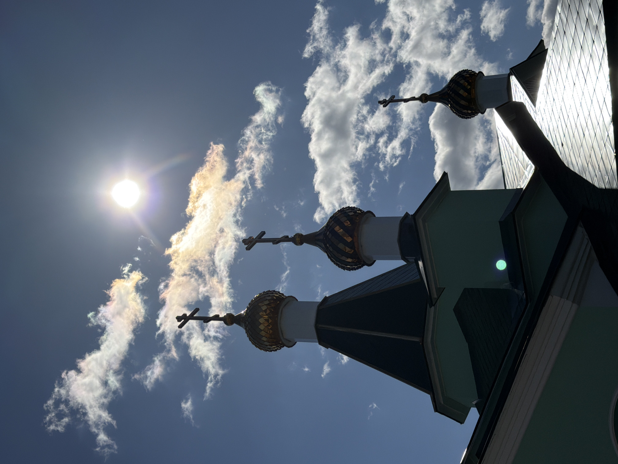 This is a low-angle, upward-facing view of a building with three domes, likely an Orthodox church, against a backdrop of a bright blue sky with scattered clouds and the sun. The domes are golden and topped with crosses. The sun is visible in the upper left portion of the image, along with a cloud that has a rainbow appearance. The building is on the right side of the image, and is partially in shadow. The location is Chaikovsky, Russia.