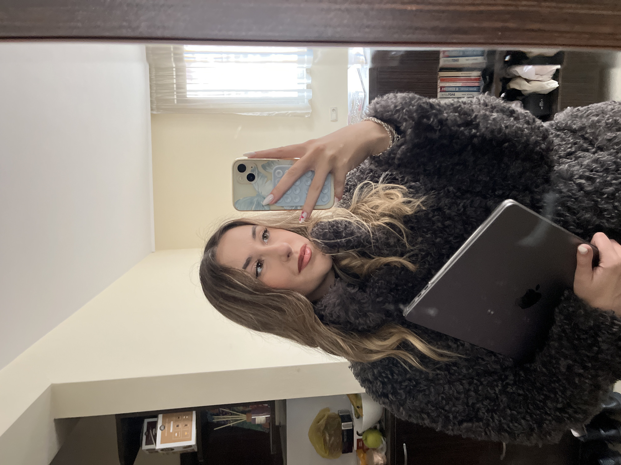 A young woman is taking a selfie in a mirror. She is wearing a fuzzy gray jacket and holding an iPhone in her left hand and a laptop in her right. Her hair is long and wavy. The mirror reflects a room with a window covered by blinds, a cabinet filled with books and other items, and a wall. I cannot pinpoint the exact location of the photo in Sofia, Bulgaria without more information.
