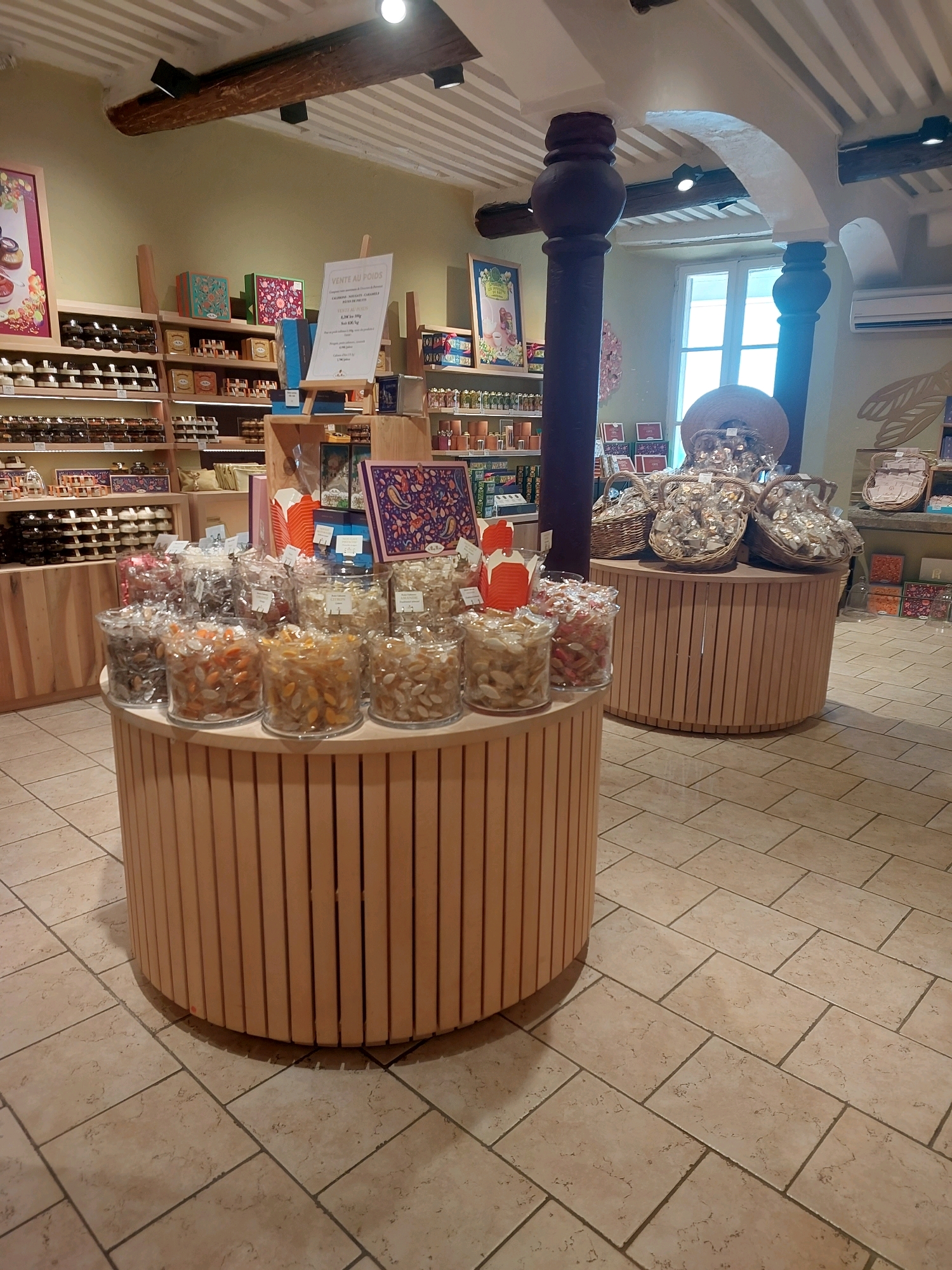 This image depicts the interior of a confectionery shop located in Saint-Paul-de-Vence, France. The shop is well-lit with natural light streaming from a large window in the background. The floor is tiled with large, beige square tiles.
The foreground is dominated by two circular wooden display units. The unit on the left is partially visible and displays several clear plastic jars filled with various sweets, including candies and possibly nougat. These jars are arranged on a circular wooden surface. The unit on the right is more fully visible and features a similar arrangement of jars filled with sweets. Behind these units, there is a tall, dark wooden pillar.
In the mid-ground, more display shelving is visible, filled with packaged goods and boxes of confectionery. To the right of the central pillar, a large round basket is visible, filled with individually wrapped sweets. Another similar basket is partially visible behind it. A sign with French text, likely indicating prices or product information, is positioned on a stand between the two main display units.
The background shows more shelving units stocked with products, a framed picture on the wall, and a portion of a window with a view to the outside. The overall impression is of a well-stocked and inviting shop specializing in sweets.