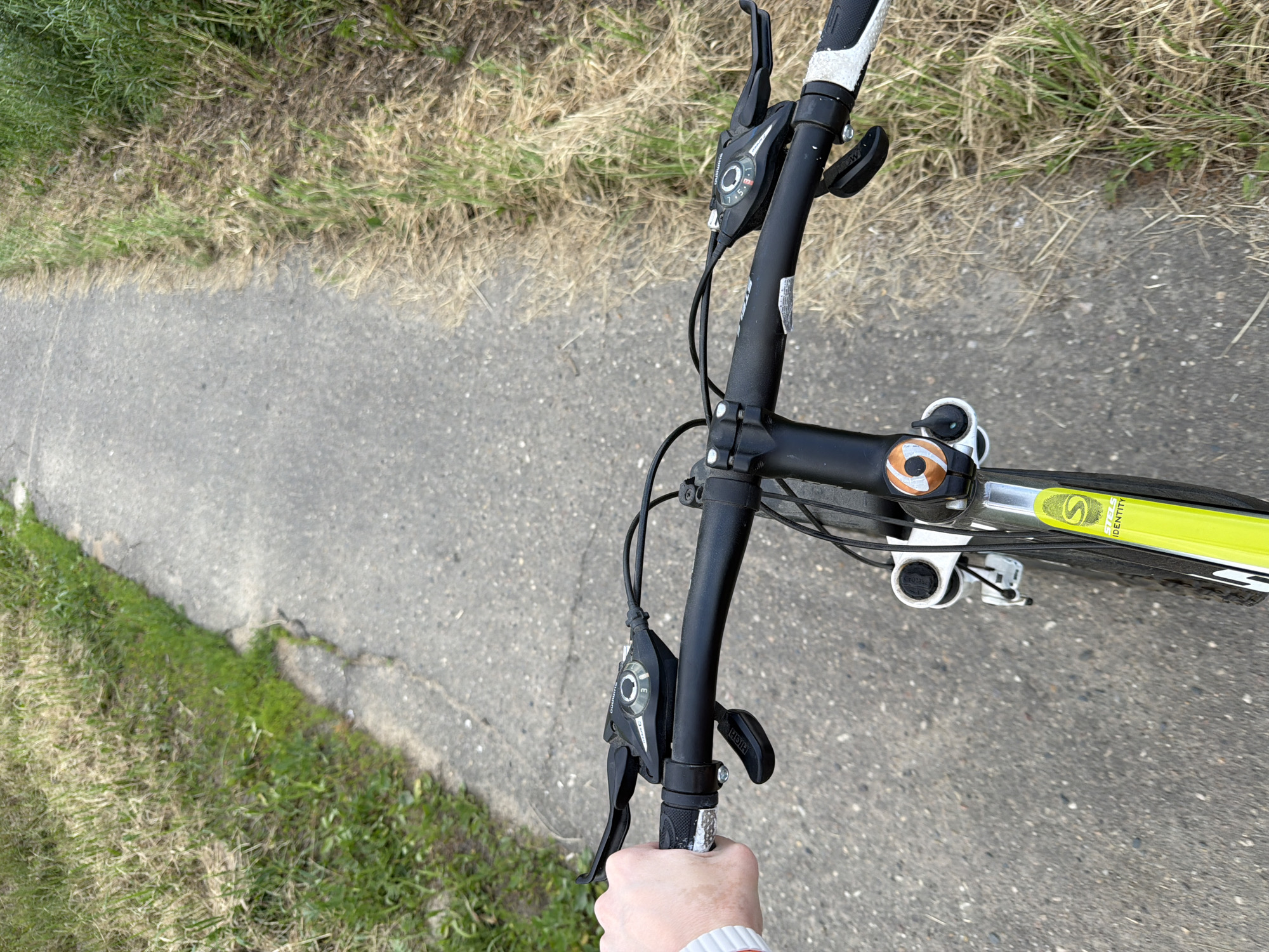 The image shows a first-person perspective of someone riding a bicycle on a paved path. The path is bordered by grass and dry vegetation on both sides. The handlebars of the bicycle are visible, with gear shifters and brake levers. A hand, with a white sleeve, is gripping the right handlebar. The bicycle has a lime green frame with some visible logos. The environment appears to be outdoors, possibly a park or a trail, in an unspecified urban or suburban location.