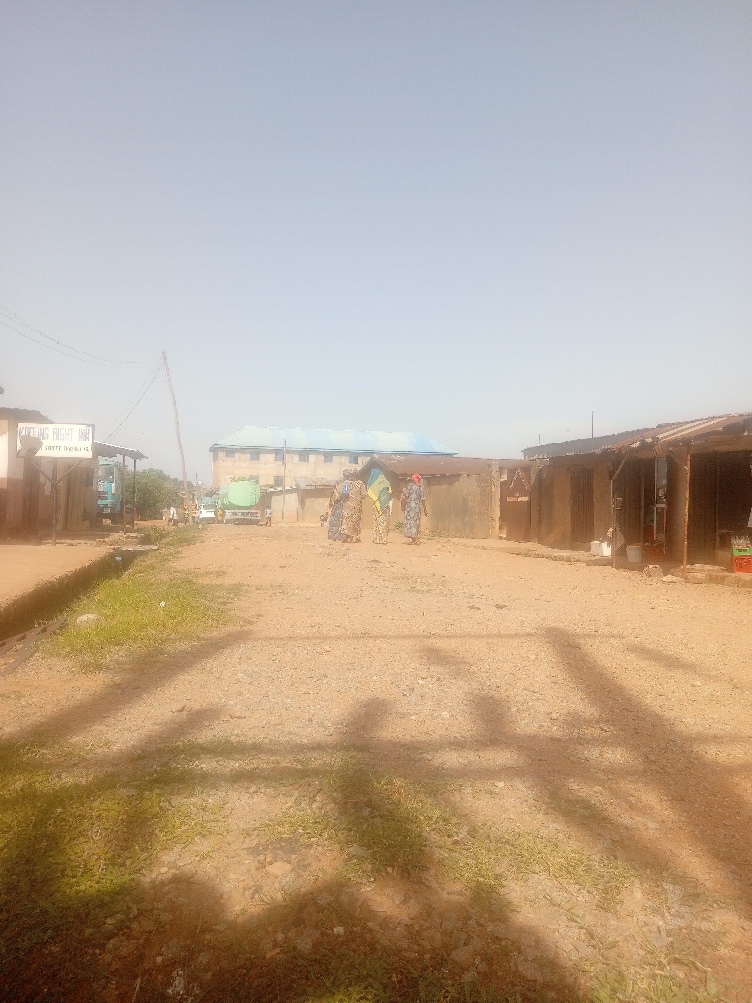 In a dusty, outdoor urban setting, a group of people are seen walking together on a dirt road. The scene appears to be in a developing area, with simple buildings and some evidence of commercial activity. A sign for "KADUNA RIGHT INN" is visible on the left, suggesting the presence of lodging. Two trucks are parked on the road, one light blue and the other green, possibly for transport or sanitation. Several individuals, including women in traditional attire, are present, some walking while others appear to be conversing. The overall impression is of a bustling, everyday scene in a Nigerian city, possibly Kaduna given the signage. The environment is characterized by a dusty atmosphere and sparse vegetation, with shadows cast across the ground, indicating it is likely daytime.